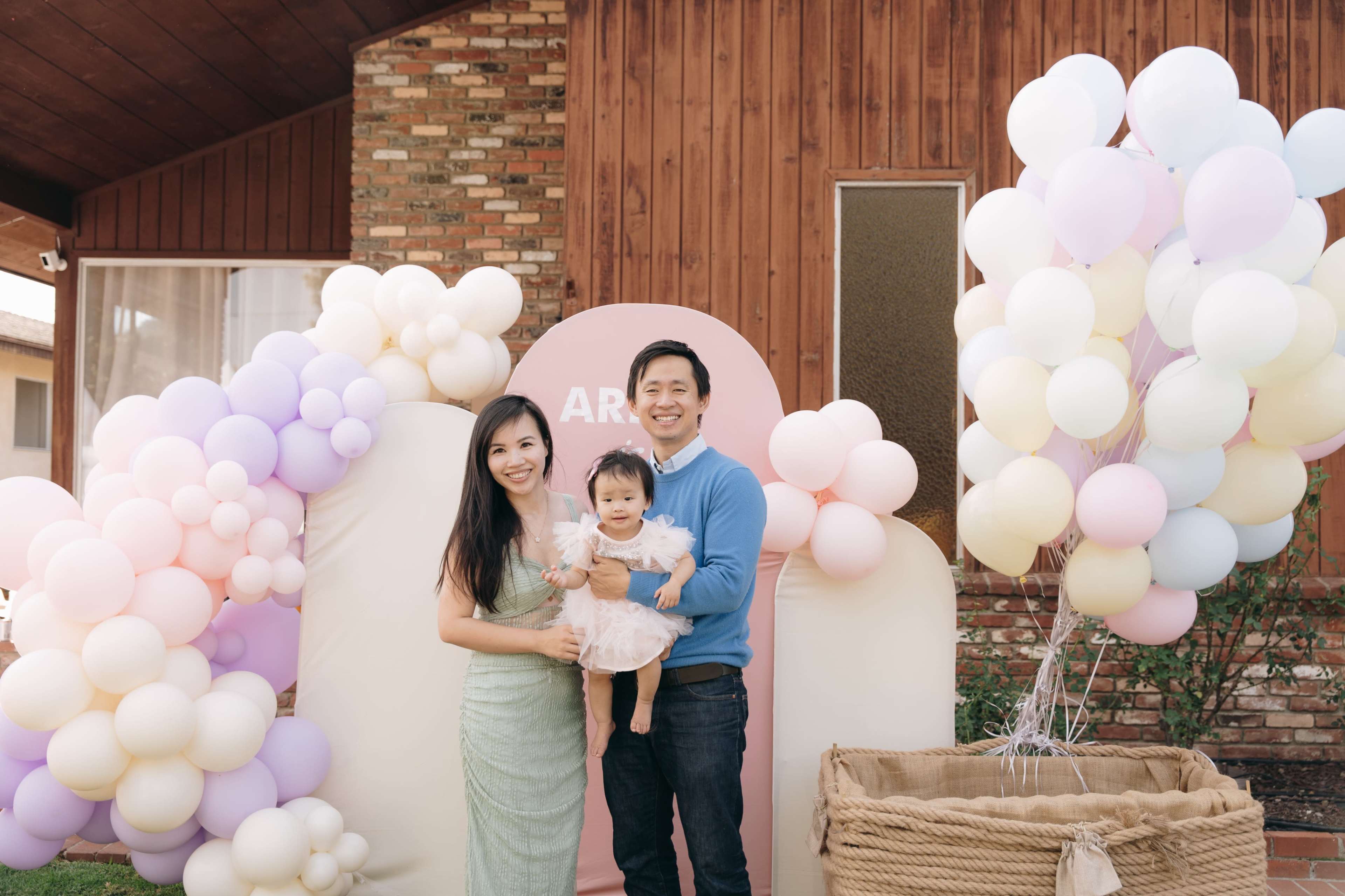 A family poses together in front of a festive balloon arch with a sign that reads "ARIEL."