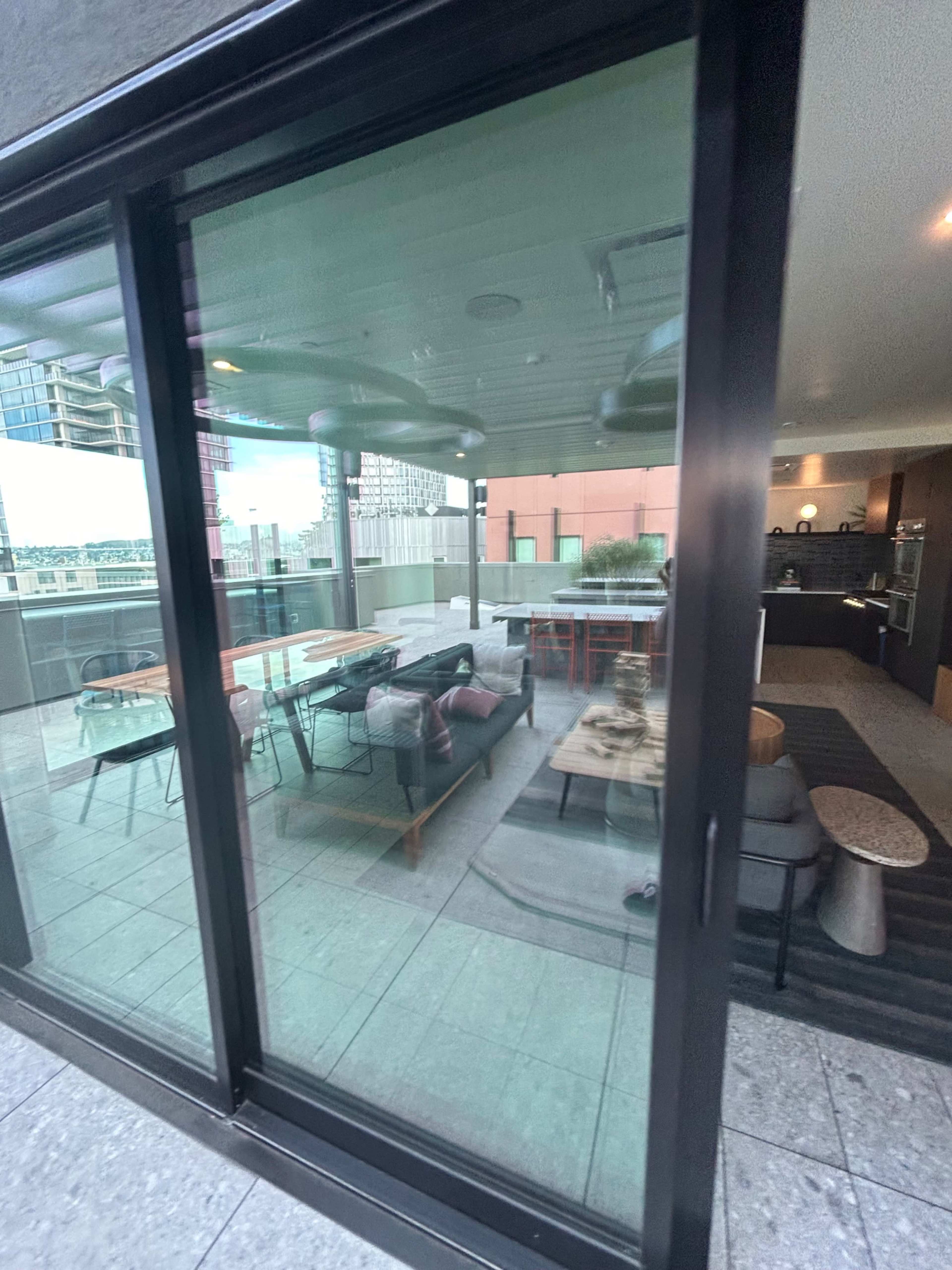 A glass door reflects a modern balcony with outdoor furniture, including a sofa and tables, overlooking a cityscape.