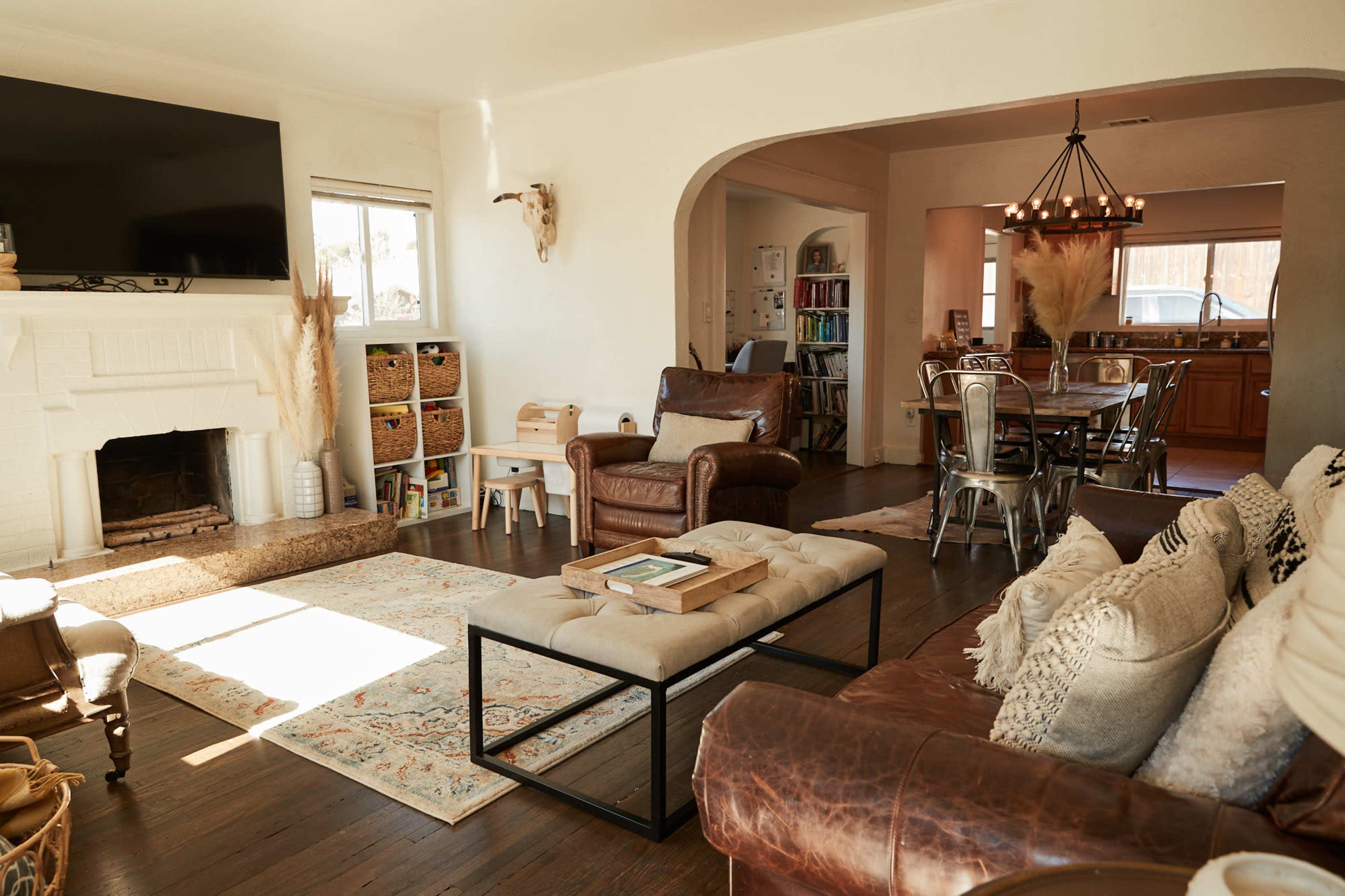 A cozy living room features a sectional couch, a coffee table, and a television above a white fireplace, with a dining area visible in the background.