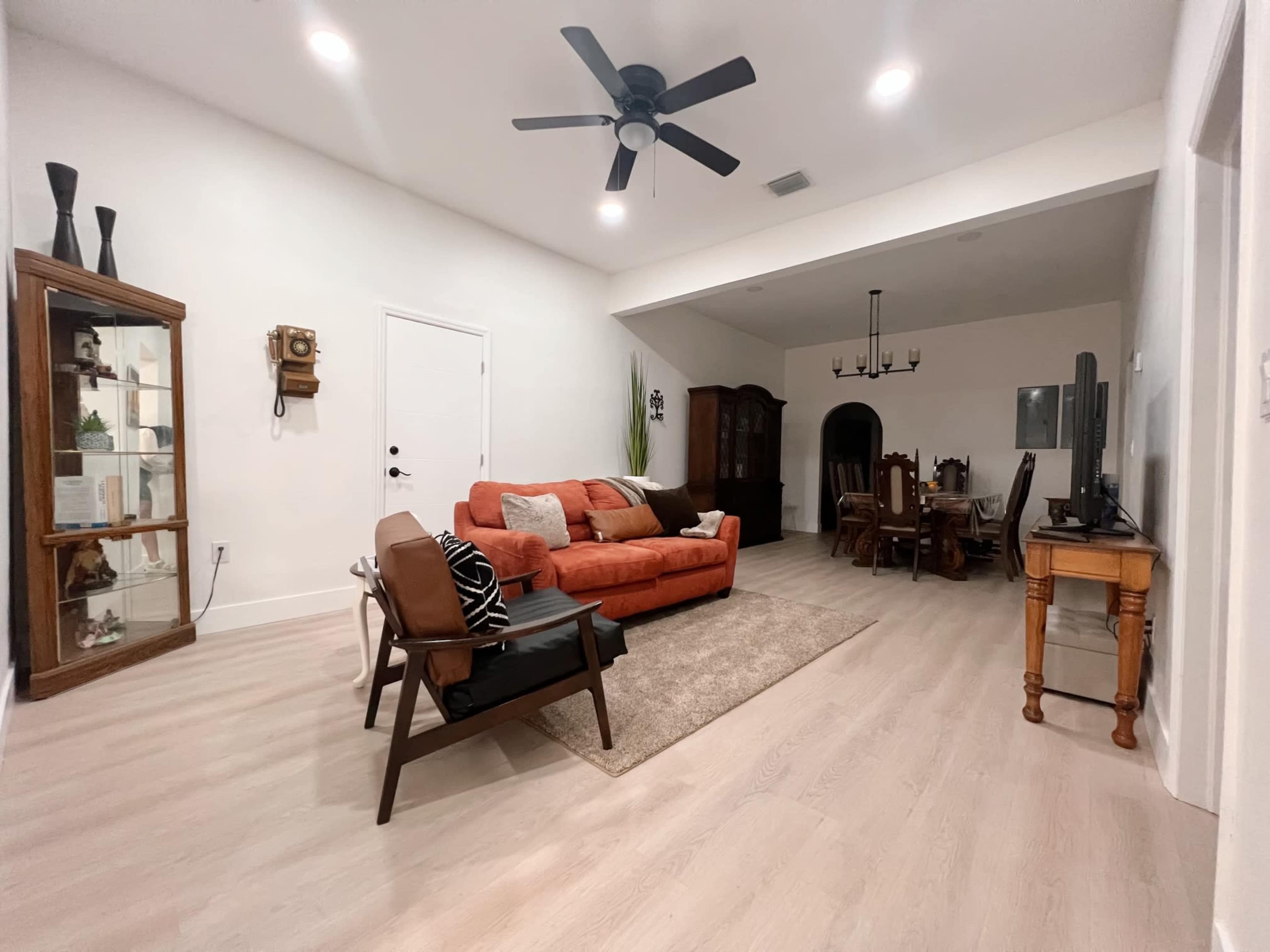 A living room with an orange sofa, a glass display cabinet, a dining area with a large table, and a ceiling fan.