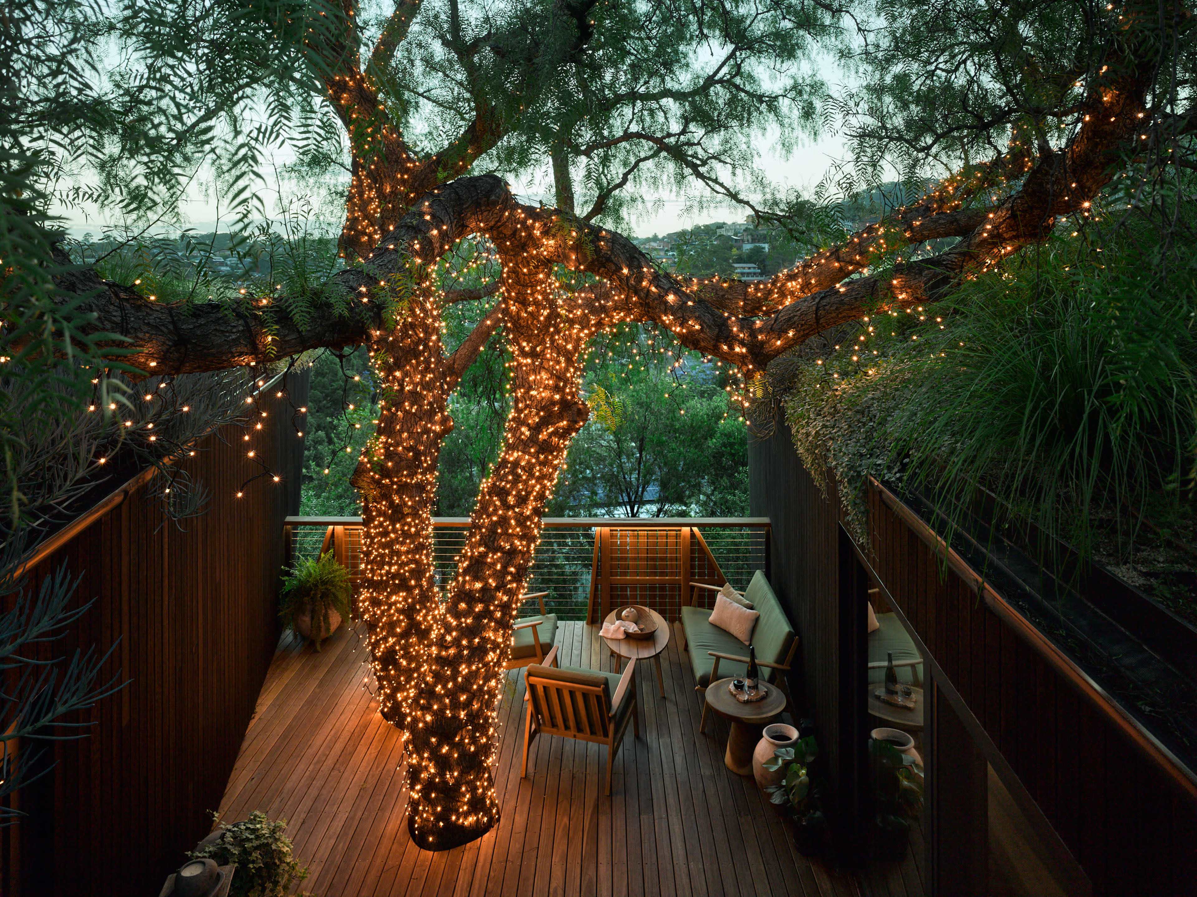 A large tree wrapped in string lights stands at the center of a wooden deck, surrounded by seating and greenery.