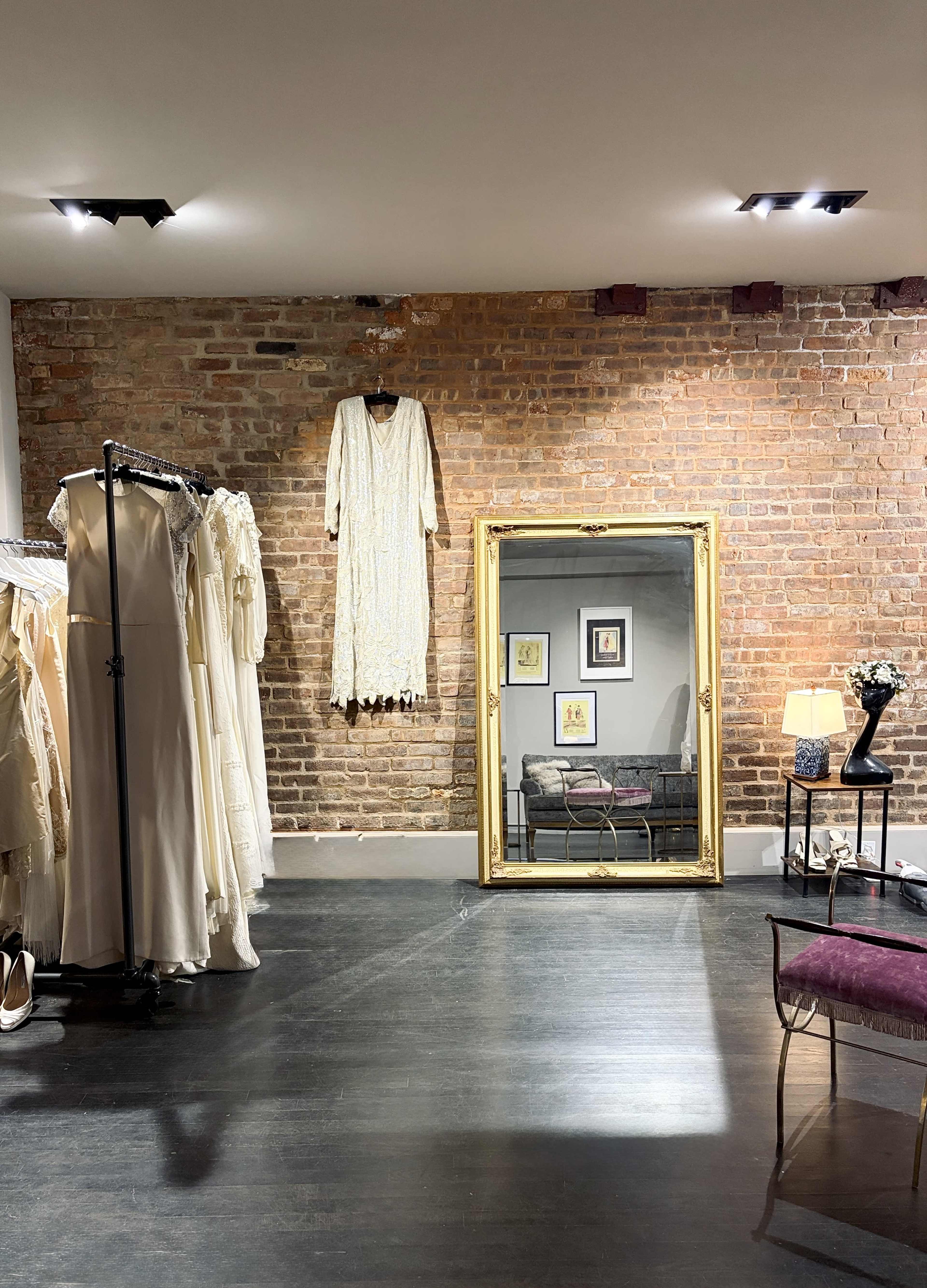 The image shows a boutique with hanging dresses on racks, a large ornate mirror, and a decorative chair next to a lamp against a brick wall.