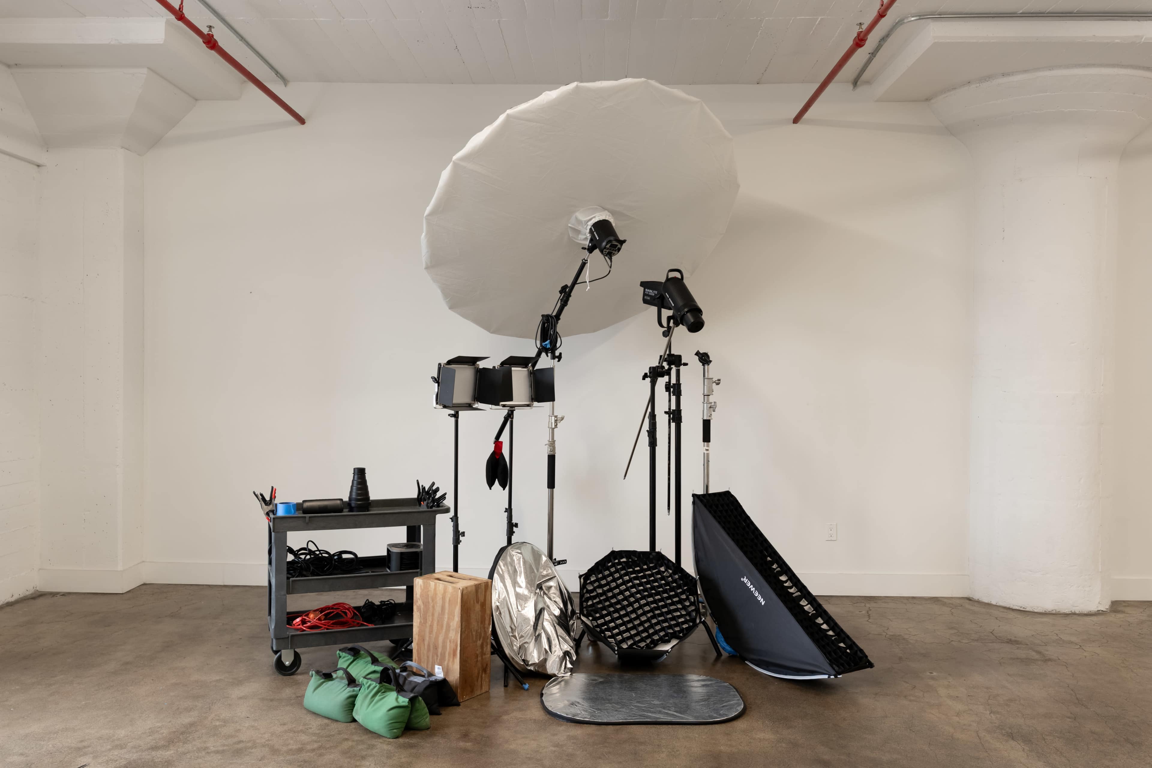 The image features various photography equipment, including light modifiers, stands, a cart with cables, and bags arranged in a minimalist studio space.