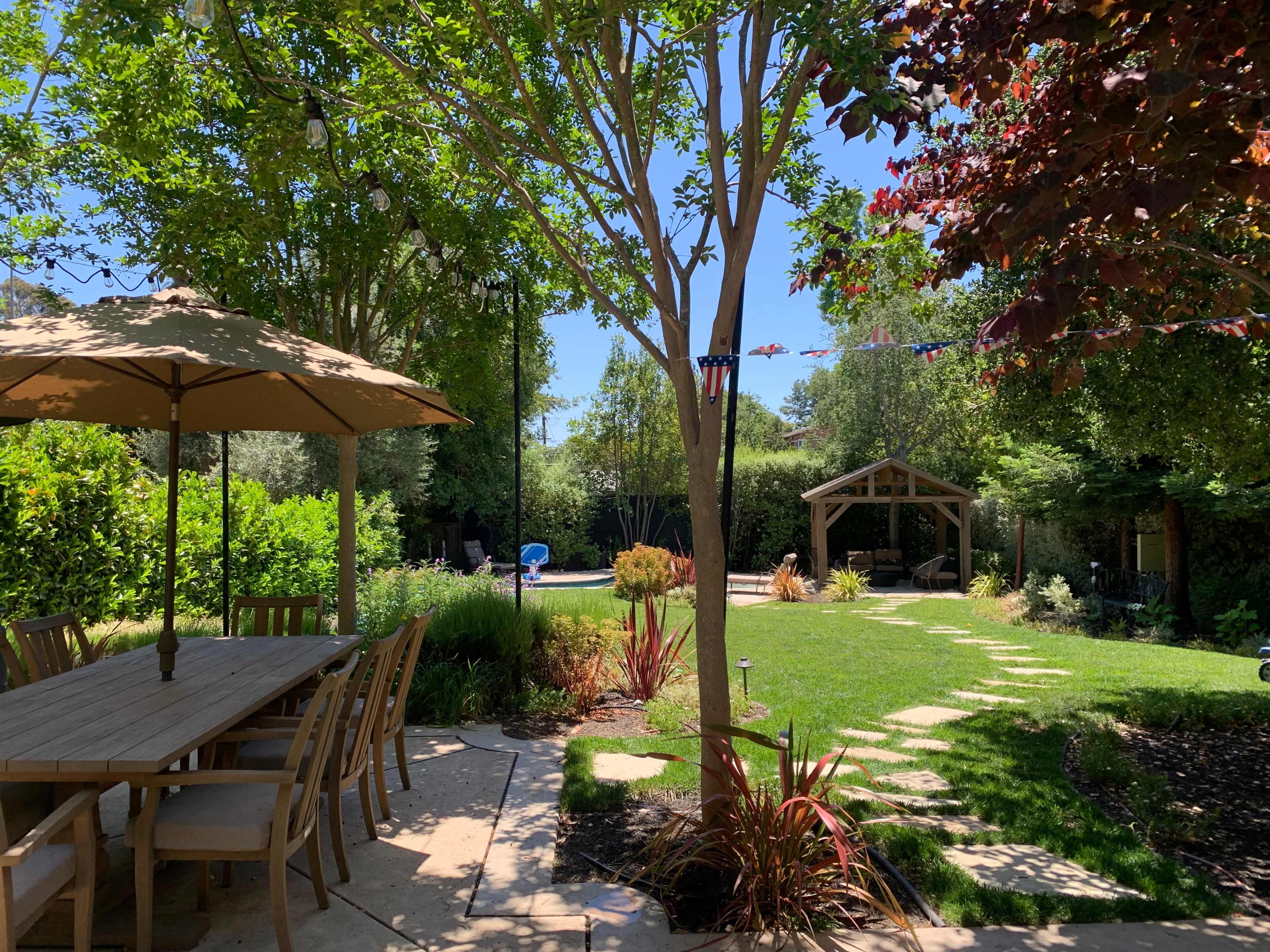 A patio area with a wooden dining table and umbrella overlooks a landscaped garden featuring a lawn, stone pathway, and a gazebo.