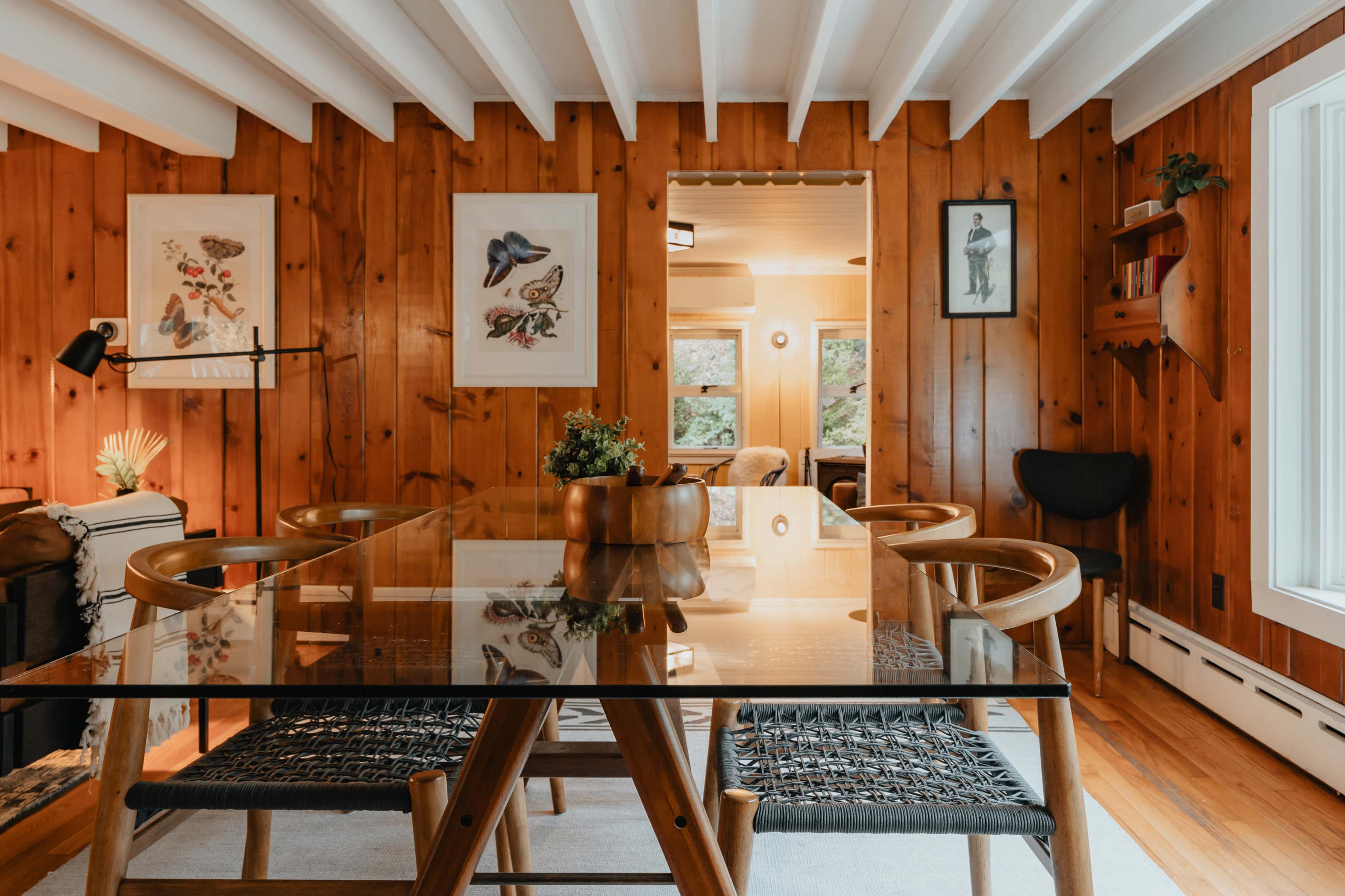 A dining area features a glass table surrounded by wooden chairs, with paneled walls and framed artwork.