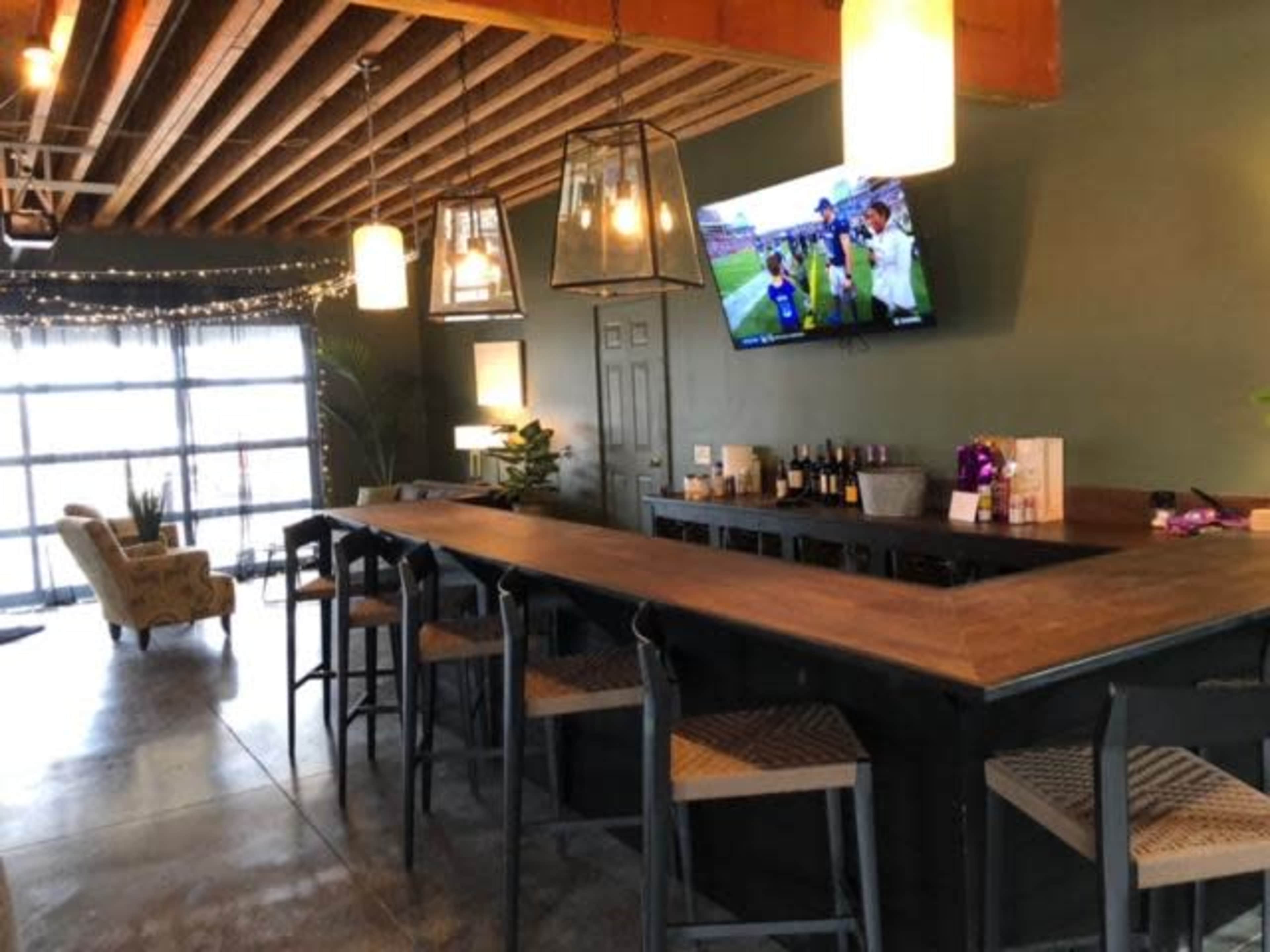 A modern bar area features a long wooden countertop, high stools, and a television displaying a sports event, with soft lighting and a lounge area in the background.