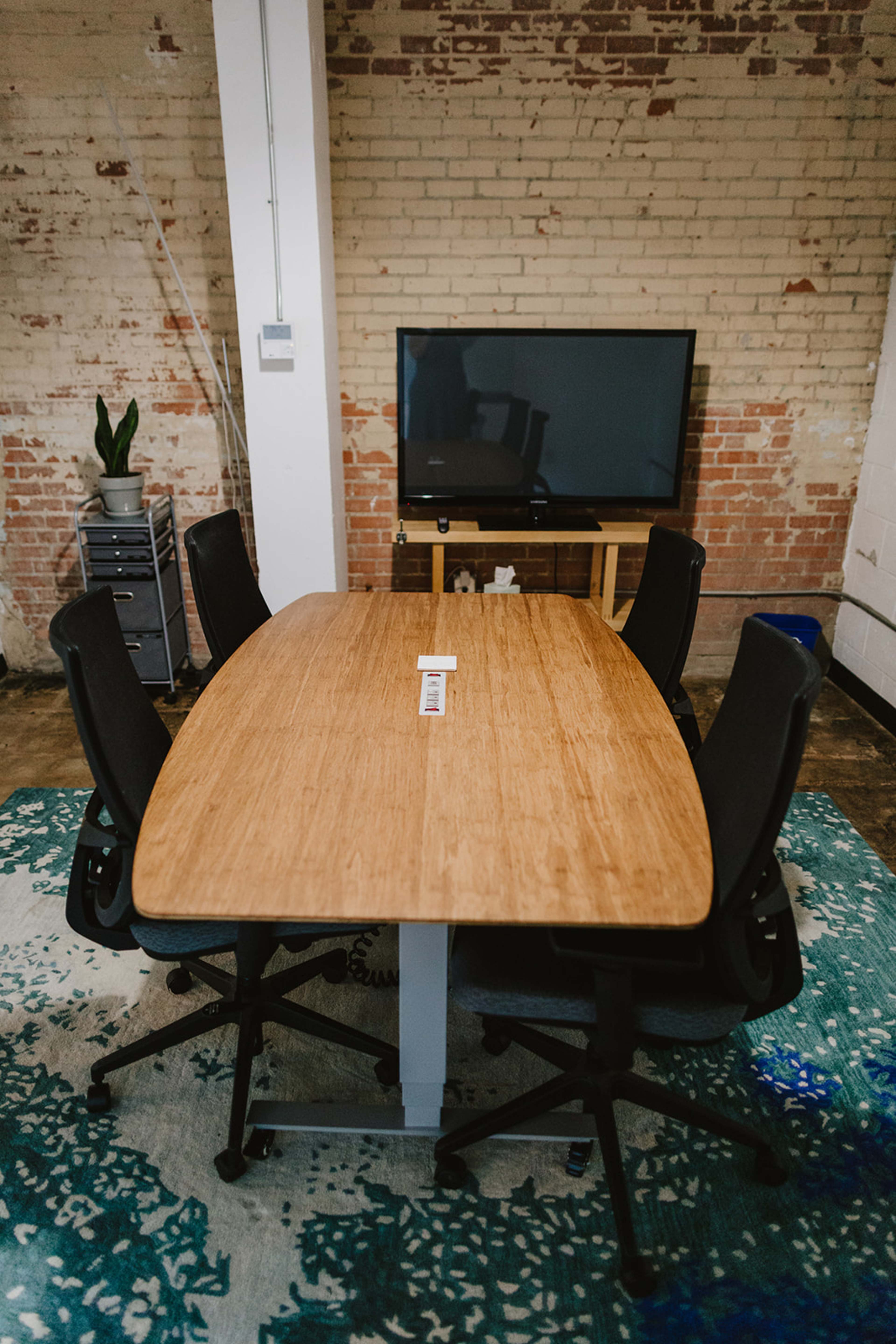 A wooden conference table with five black chairs is positioned in front of a television screen in a room with exposed brick walls.