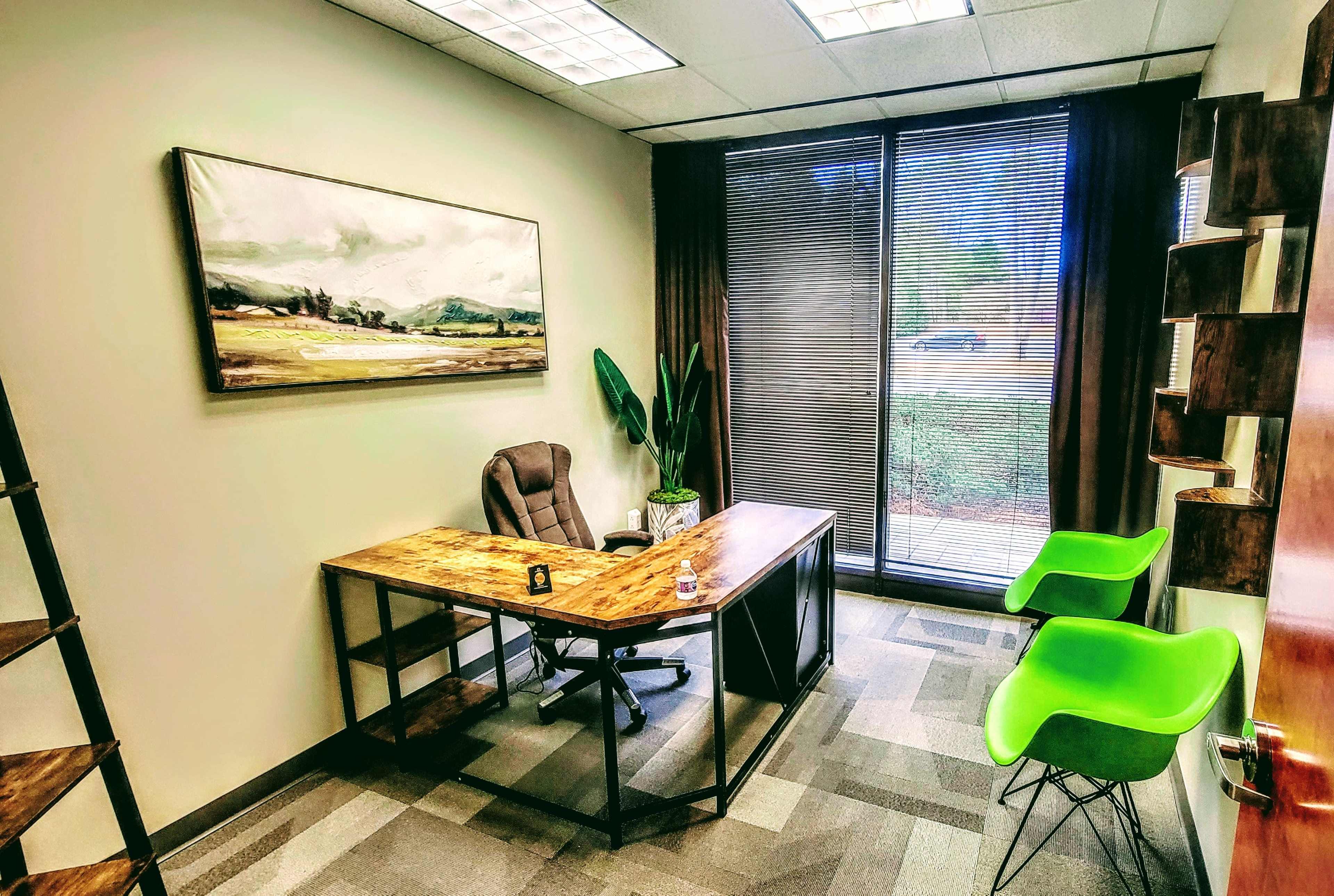 A modern office space with a wooden desk, an ergonomic chair, and two green chairs beside a large window with blinds.