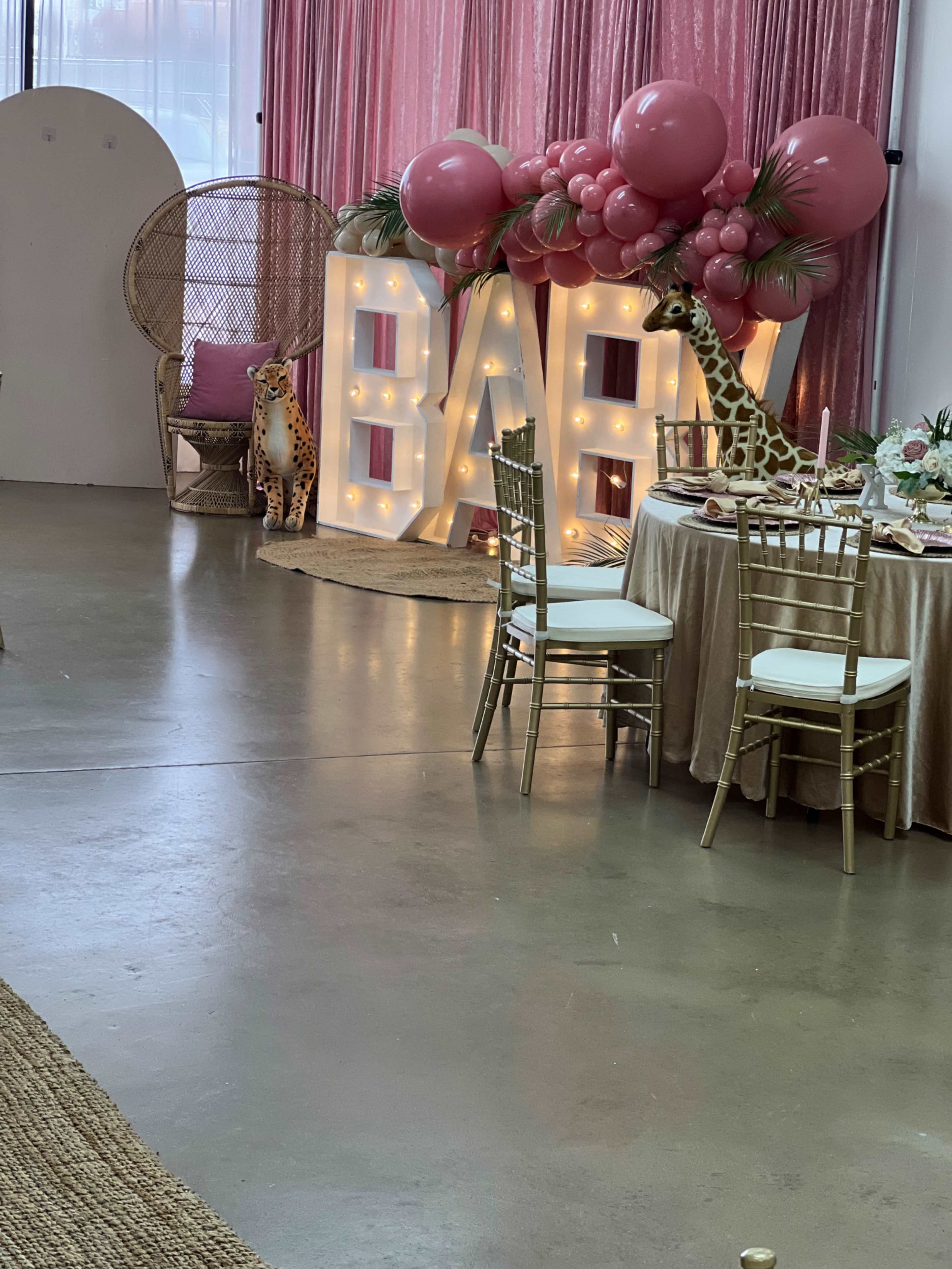 A decorated event space featuring a large illuminated "BABY" sign, a seated giraffe decoration, and a table set for a celebration.