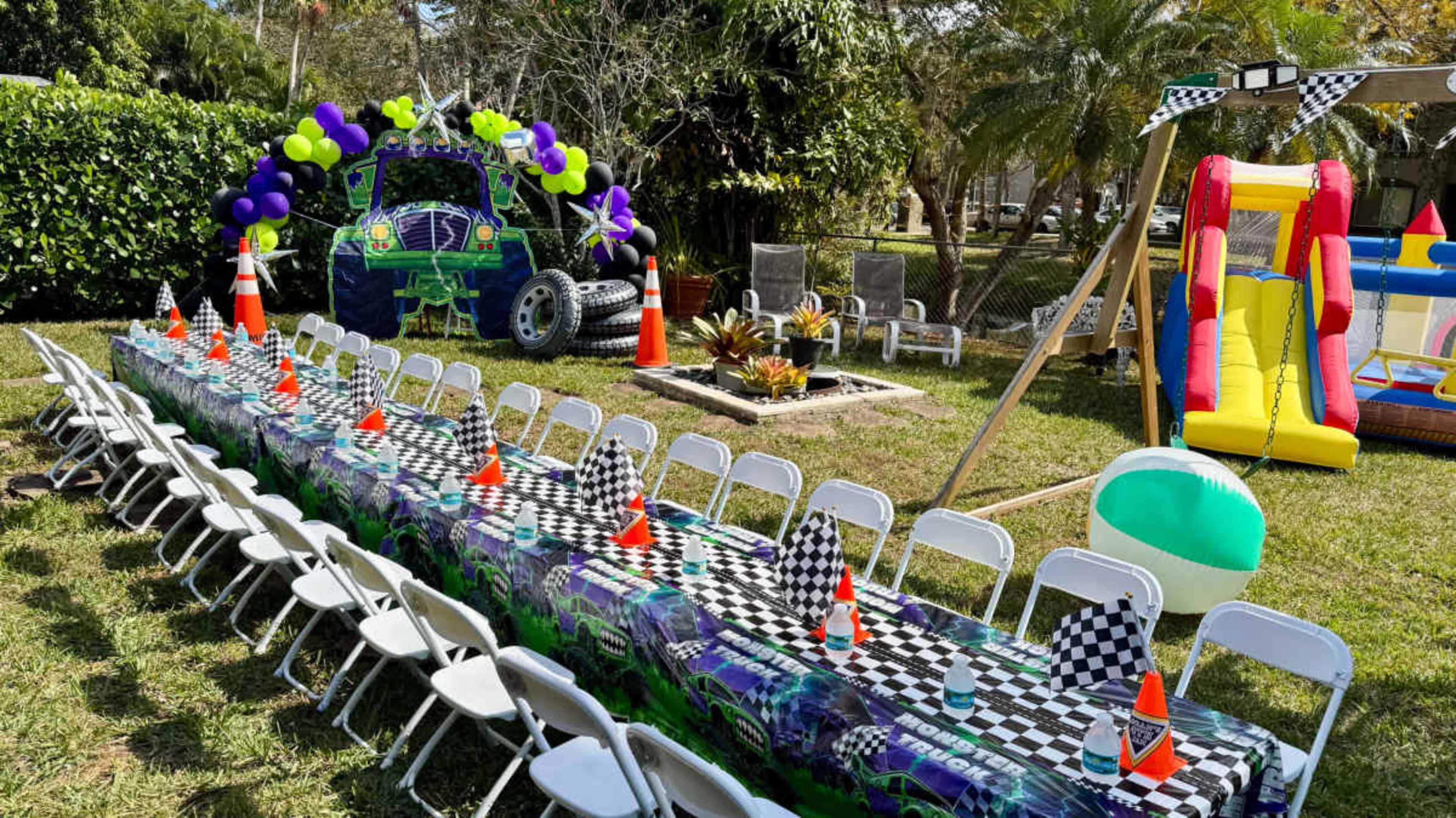 A large rectangular table is set up outdoors with checkered tablecloths, surrounded by white chairs, alongside a colorful bouncy castle, traffic cones, and decorative balloons.