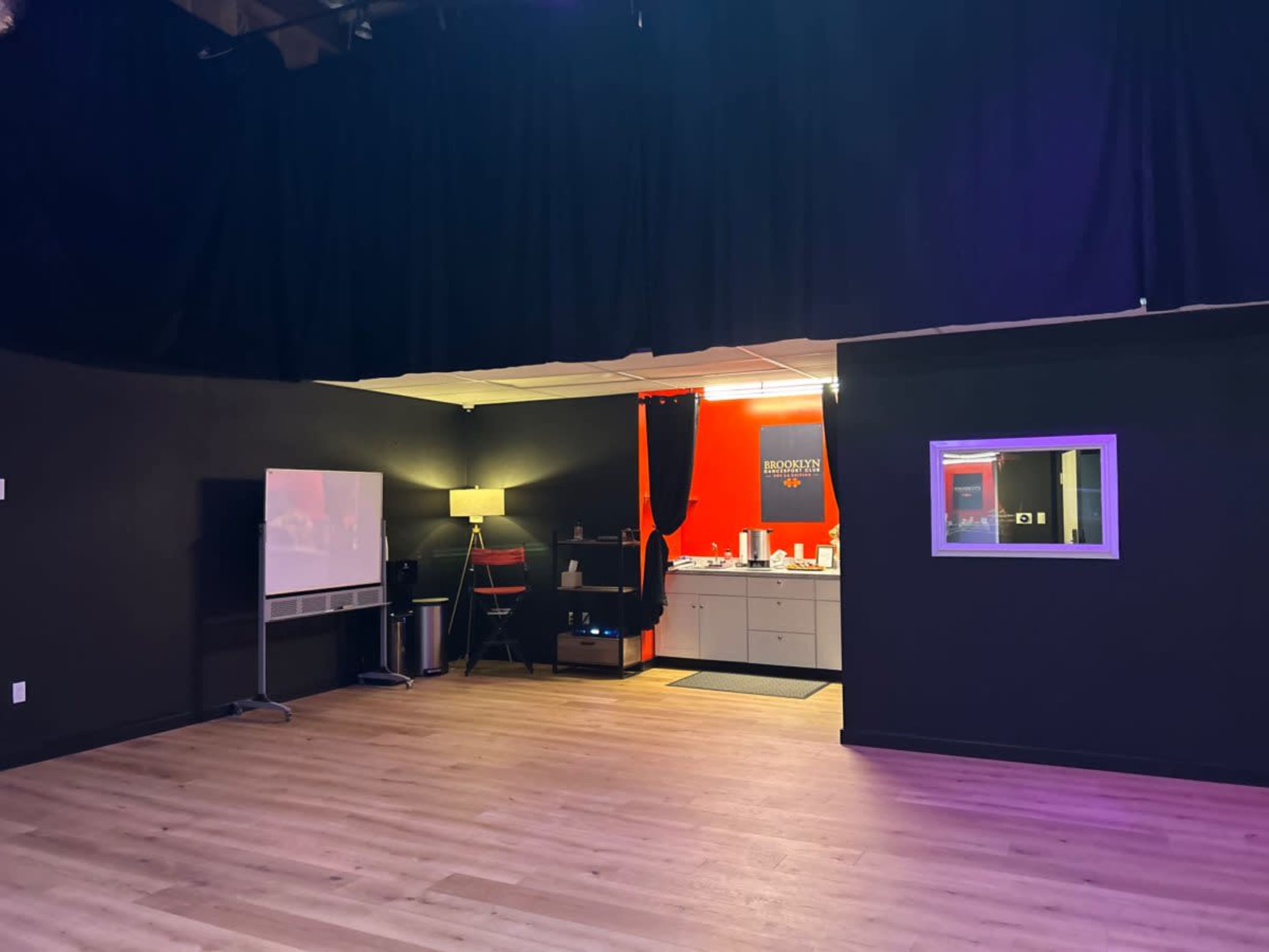 The image shows a spacious, dimly lit room with a small kitchenette at the back, separated by a black curtain, and wooden flooring.