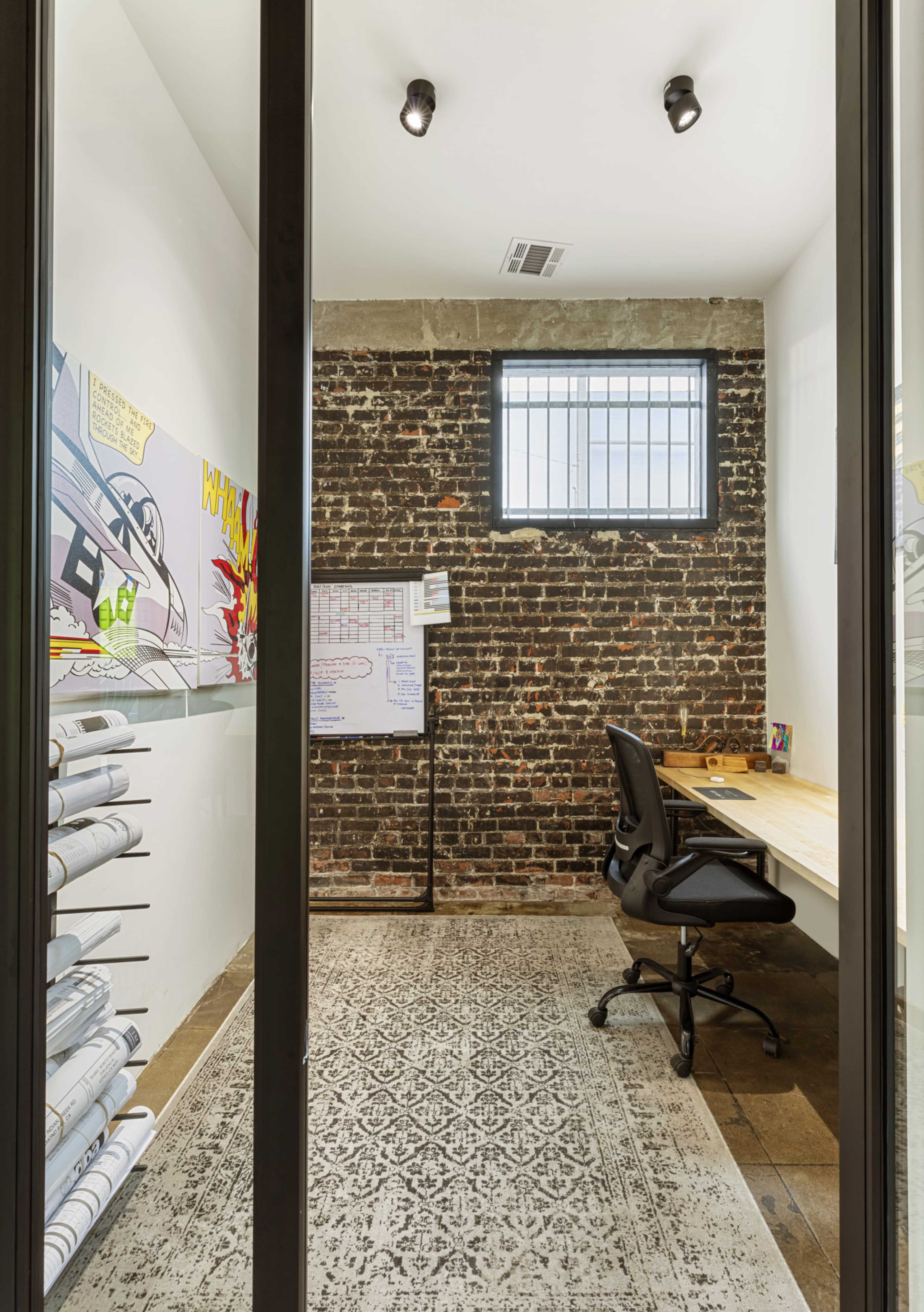 The image shows a small office with a brick wall, a desk with a chair, and rolls of paper stored on a shelf.