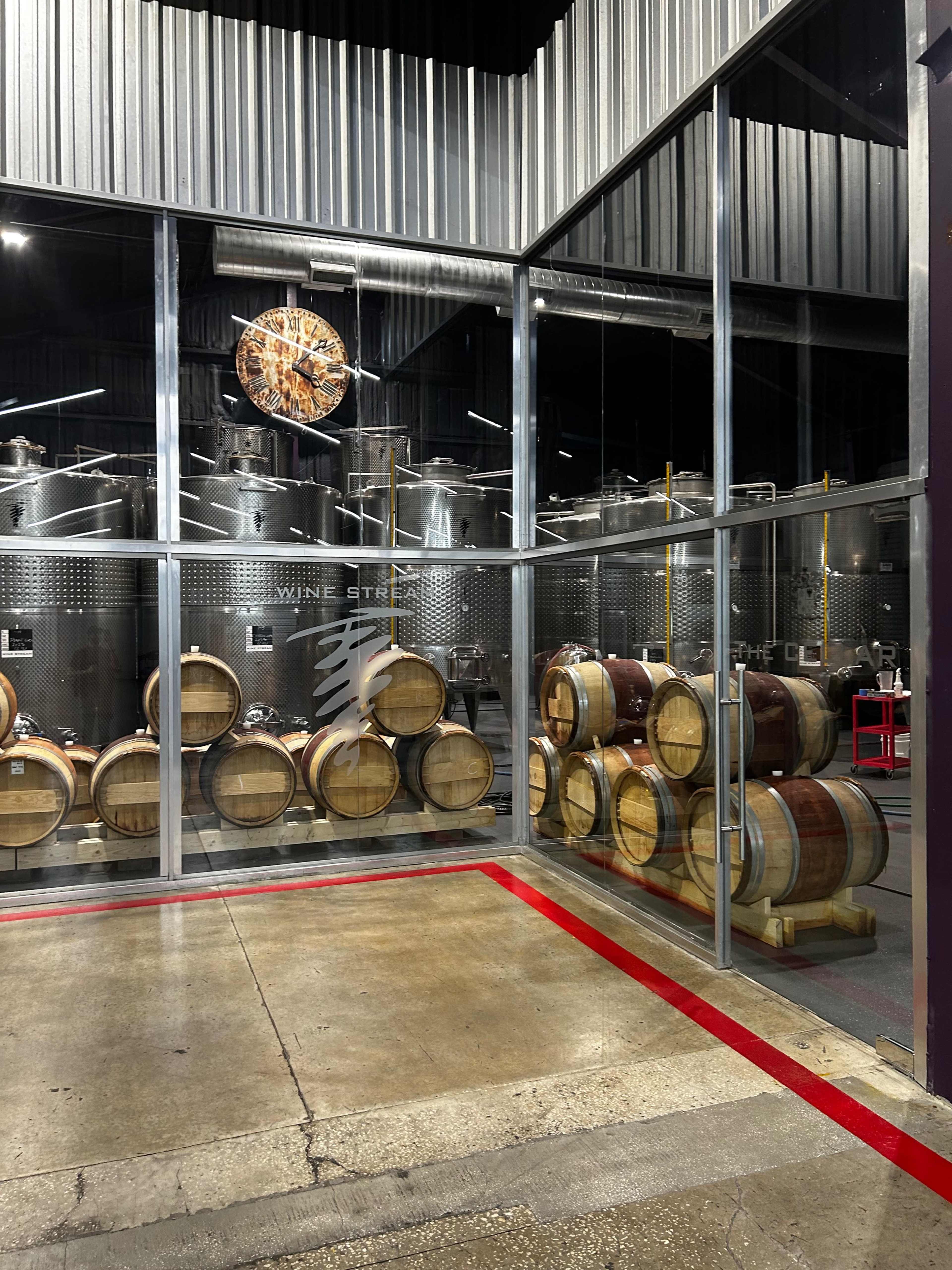 The image shows a glass-enclosed room with stacked wine barrels and stainless steel fermentation tanks in the background.
