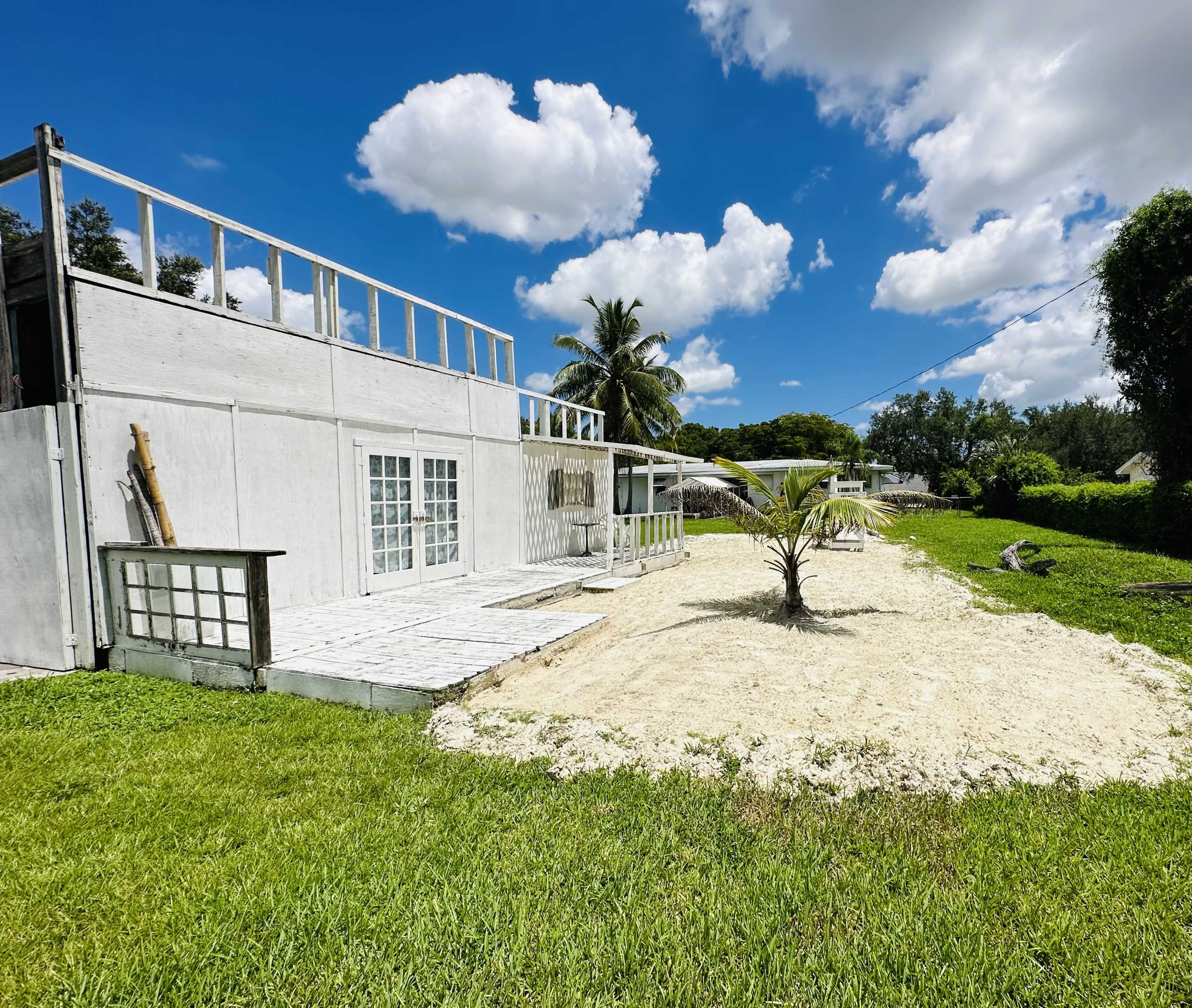 A white building with large windows sits beside a sandy area and green grass under a cloudy blue sky.
