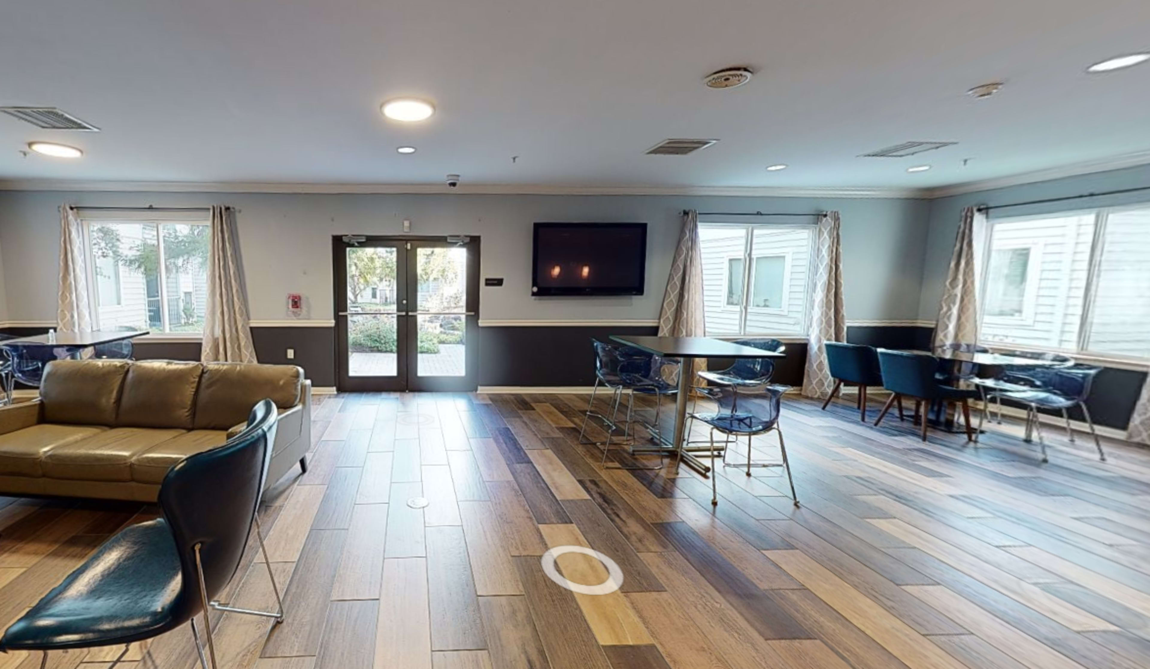 The image shows a common room with a wooden floor, couches, tables, and large windows.