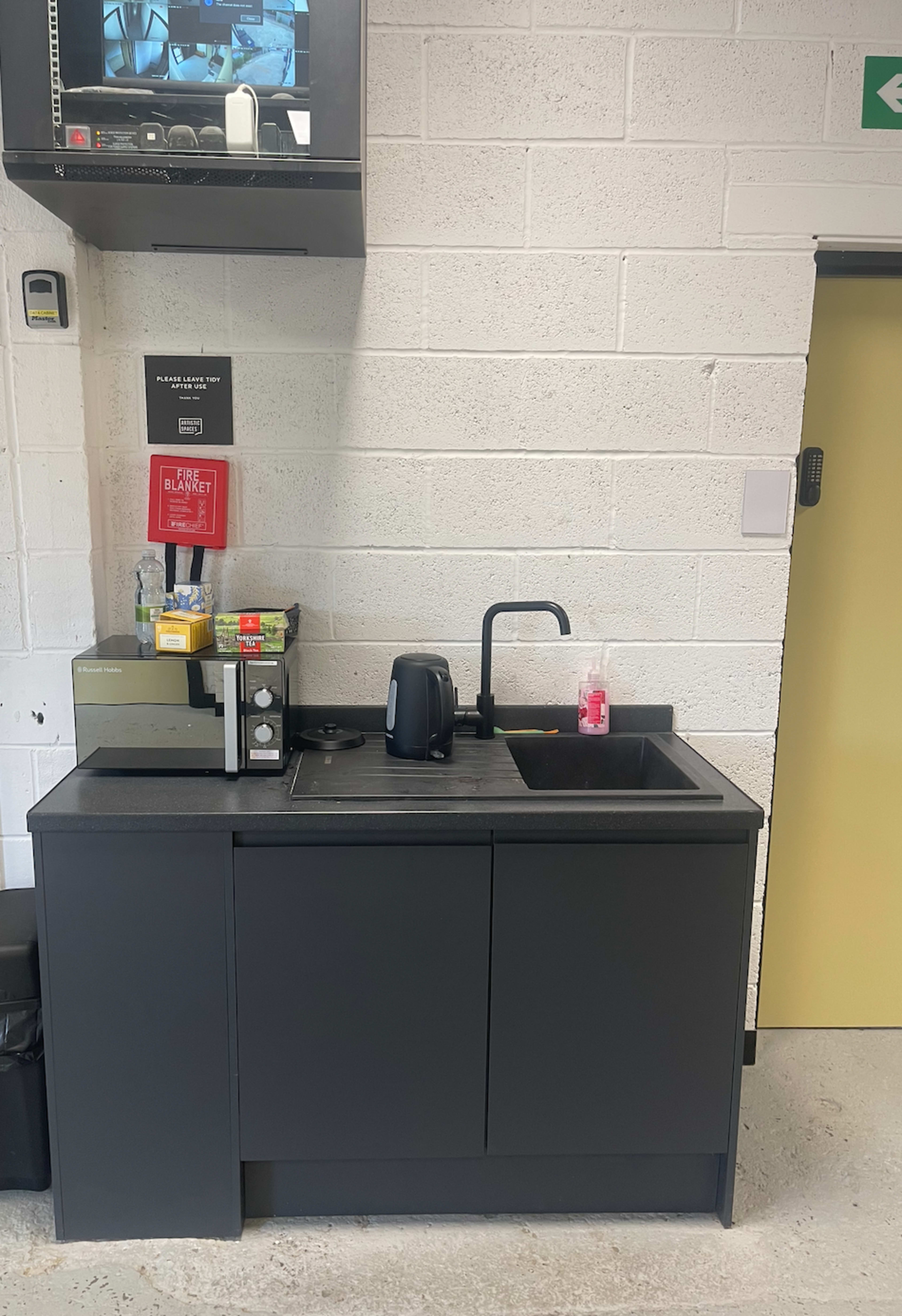 A black kitchenette with a sink, a kettle, a microwave, and various items arranged on the counter in a room with white brick walls.