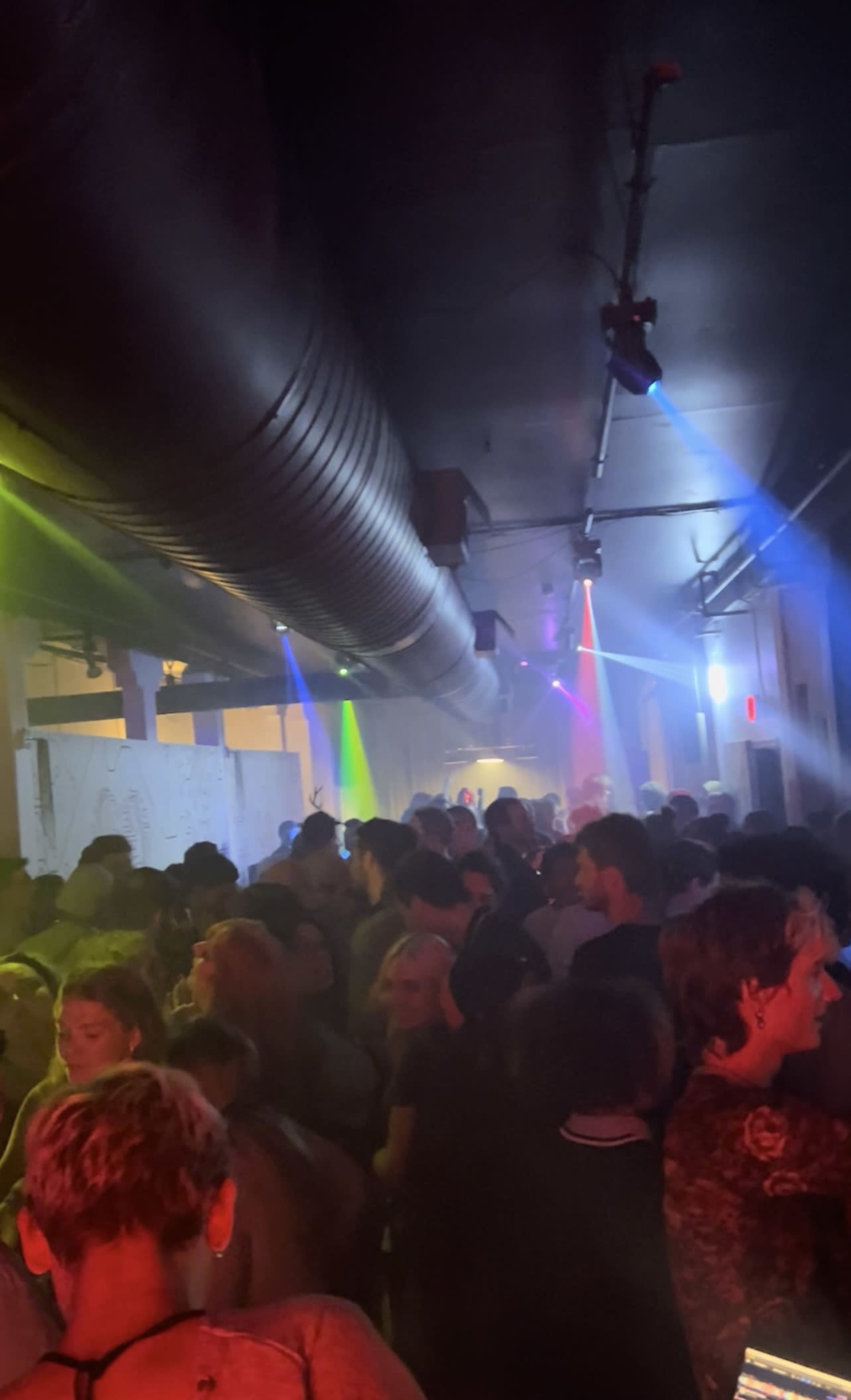A crowded indoor venue is filled with people dancing under colorful lights and a haze of smoke.