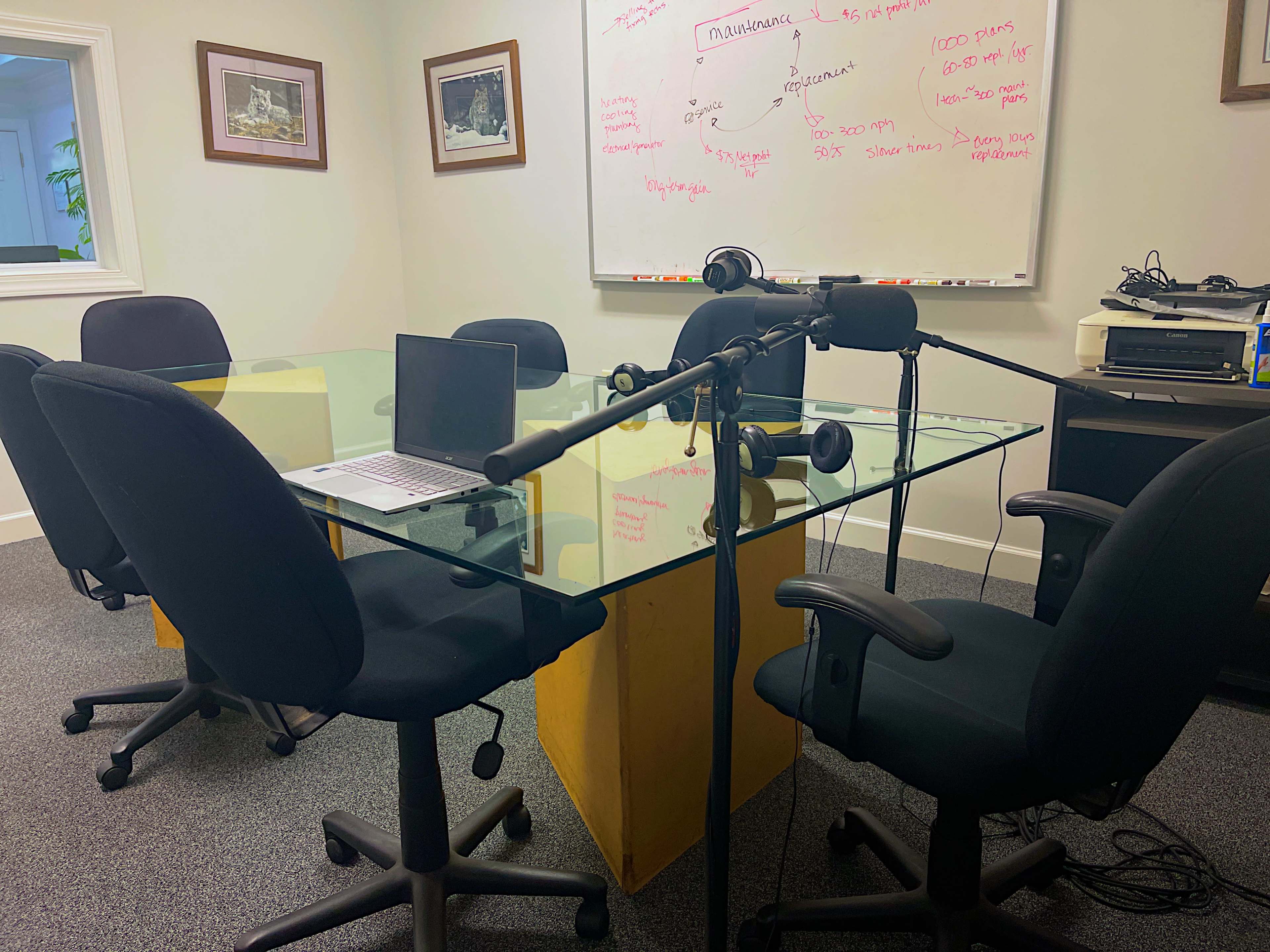 The image shows a conference room with a glass table surrounded by six black chairs, a laptop, and a microphone set up on a stand near a whiteboard covered in handwritten notes.