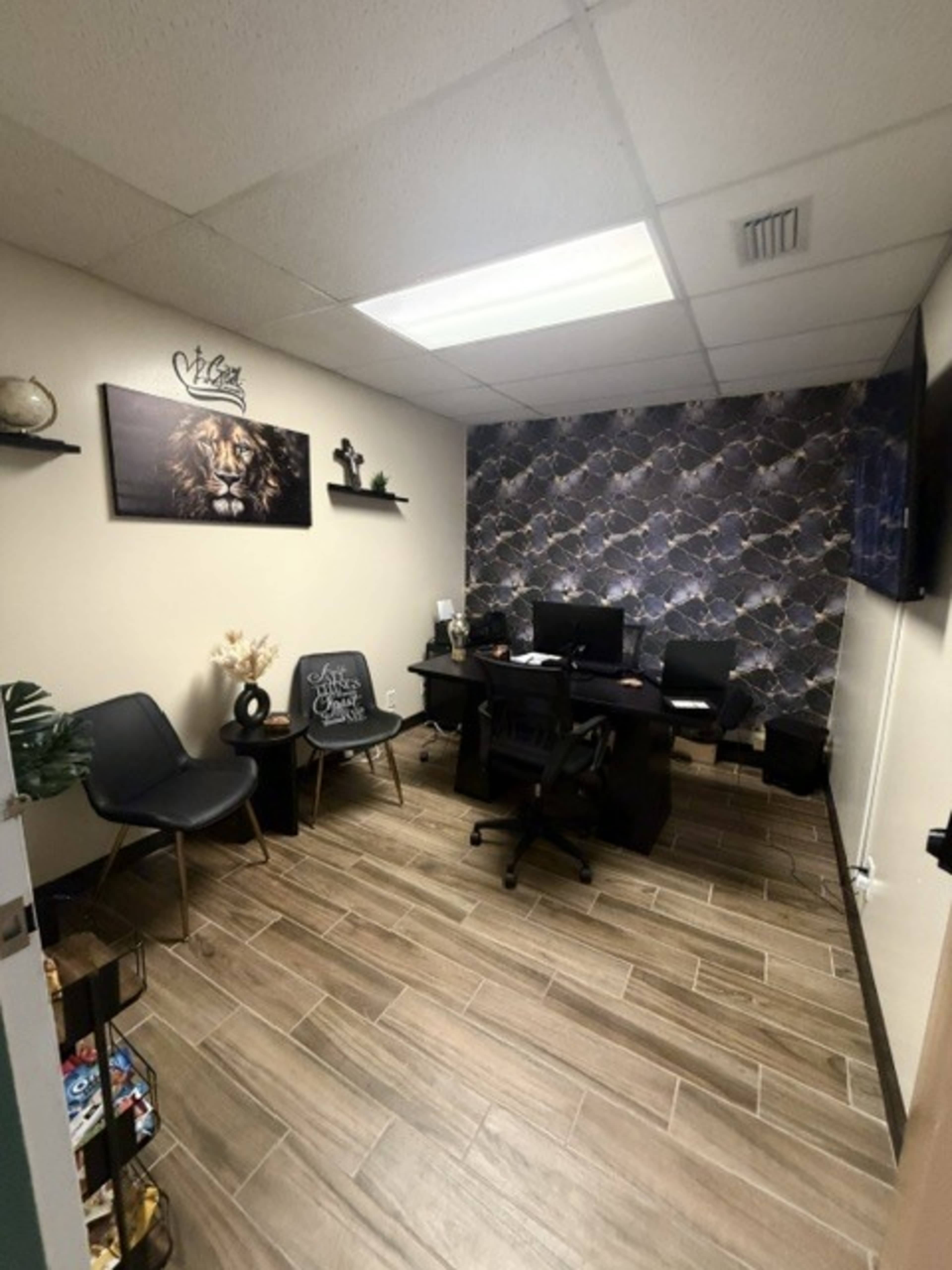 The image shows a small office with a dark patterned accent wall, two black chairs, a desk with a chair, and shelves displaying decorative items.