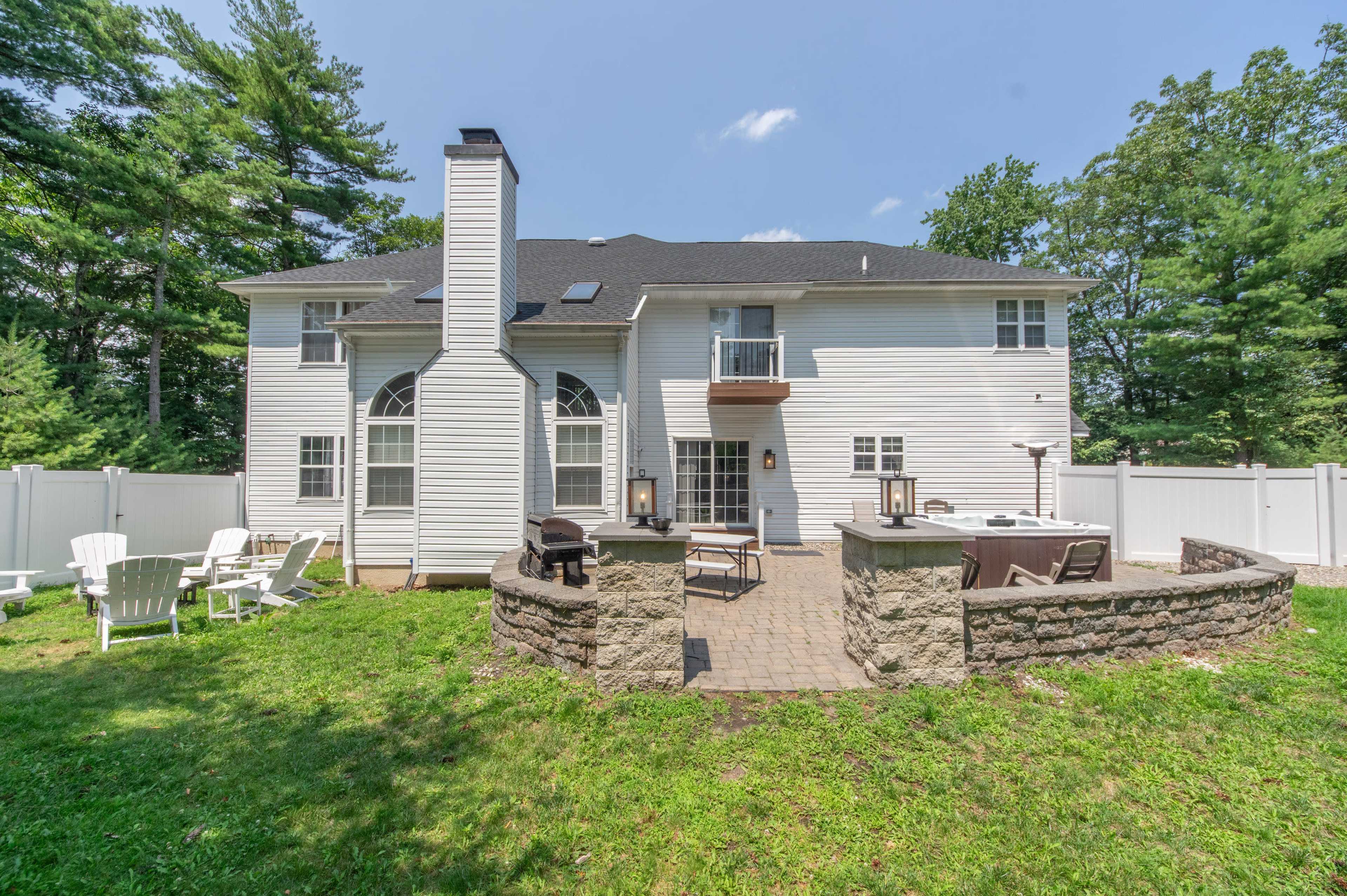 The backyard of a two-story house with a stone patio, outdoor seating, and a grassy area surrounded by trees.