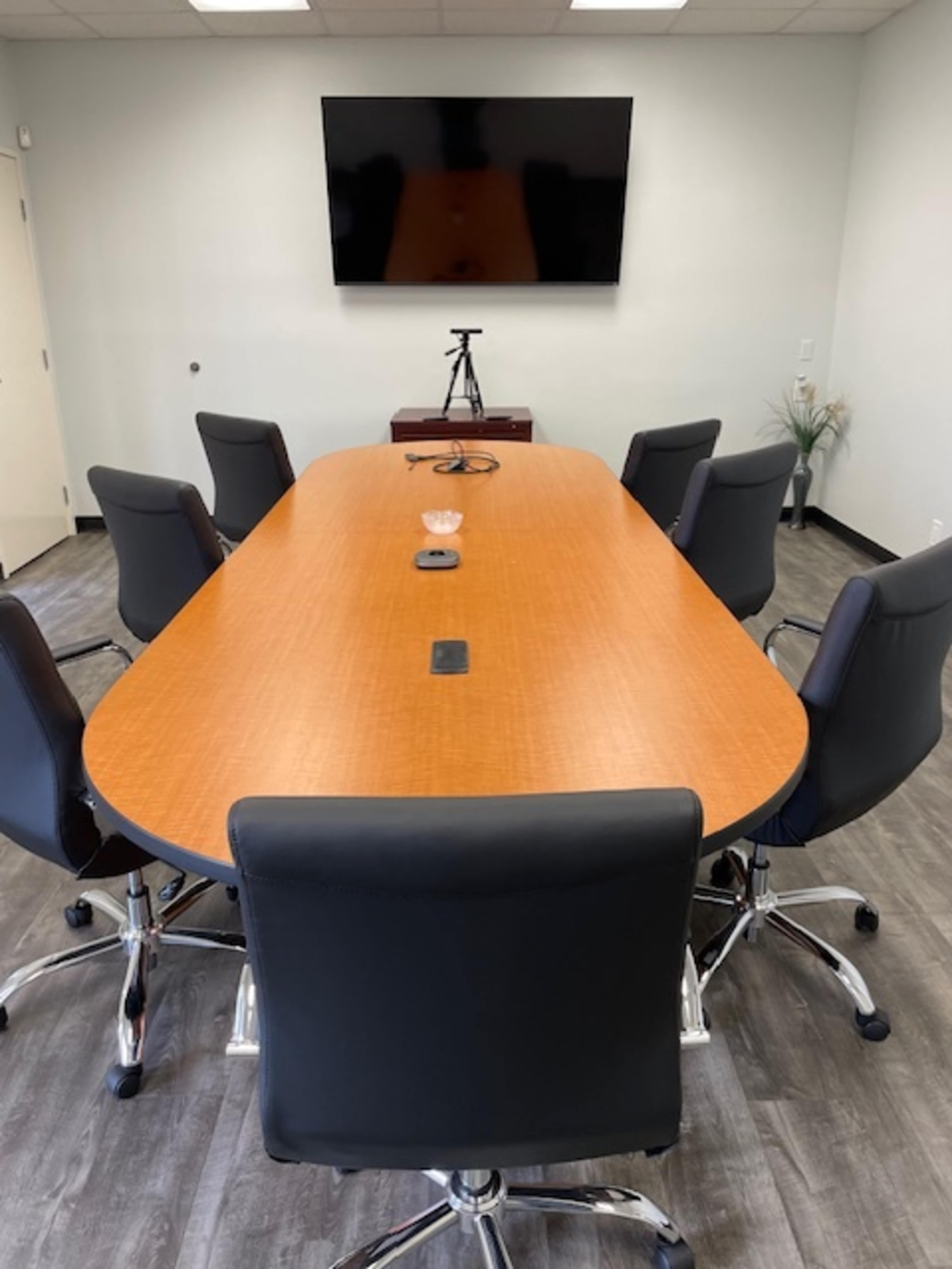 A modern conference room features a large wooden table surrounded by black swivel chairs, with a wall-mounted television and a tripod in the background.