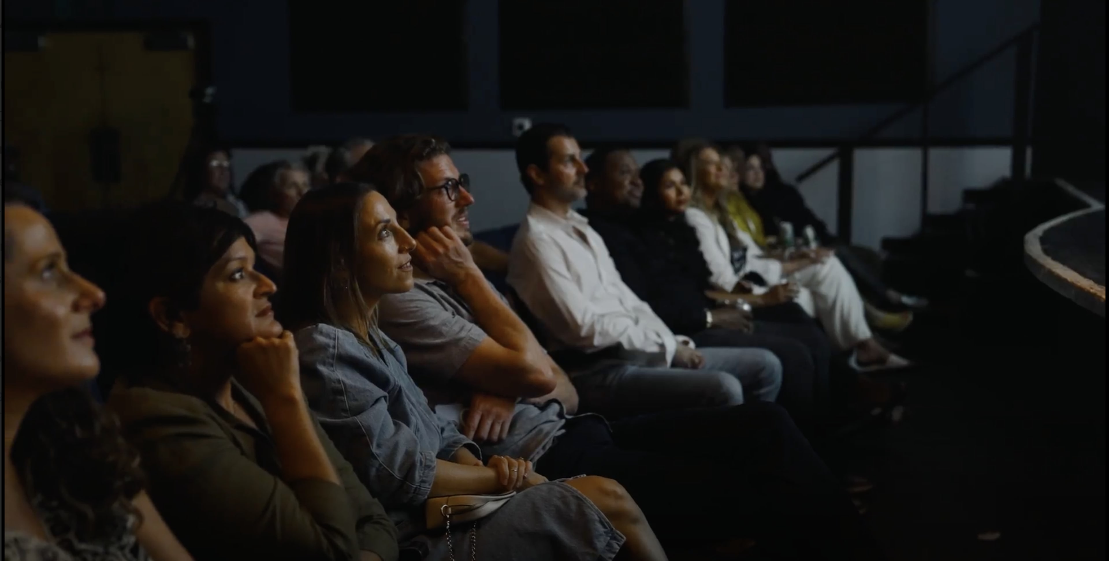 A group of people sits attentively in a darkened theater, facing a stage.