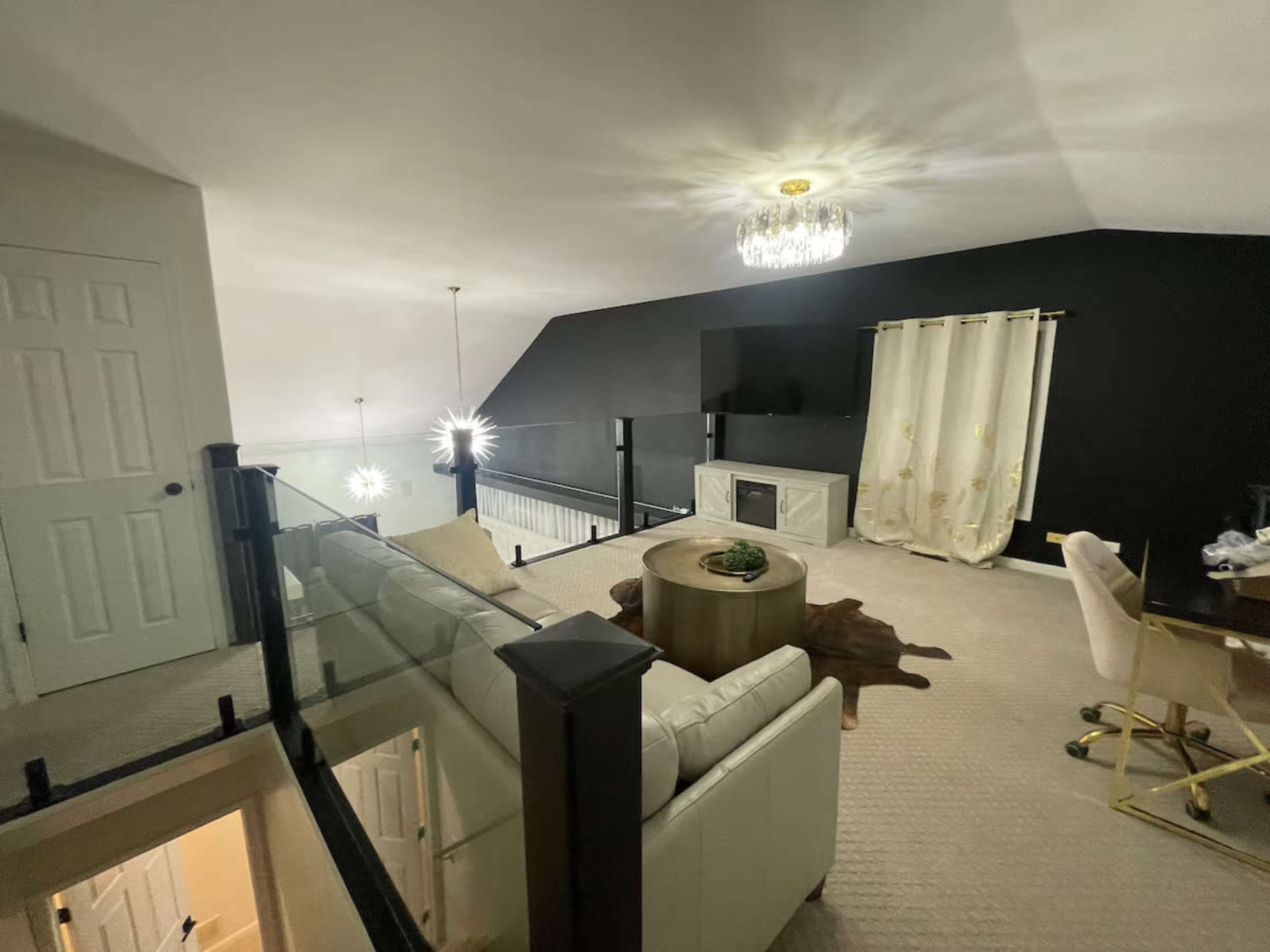 The image shows a modern loft space with a glass railing, a white sofa, a round coffee table, and a television against a black wall, complemented by a chandelier and decorative curtains.