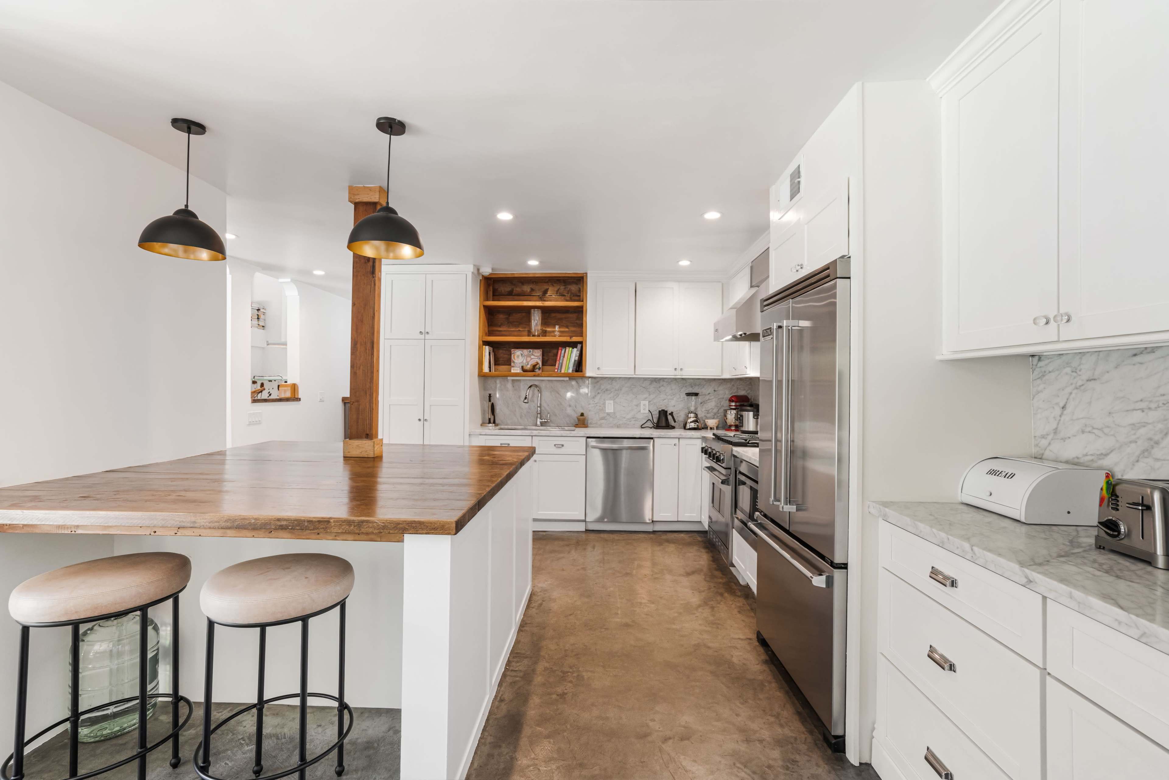 The kitchen features white cabinetry, a marble countertop, stainless steel appliances, and a wooden bar with two metal stools.
