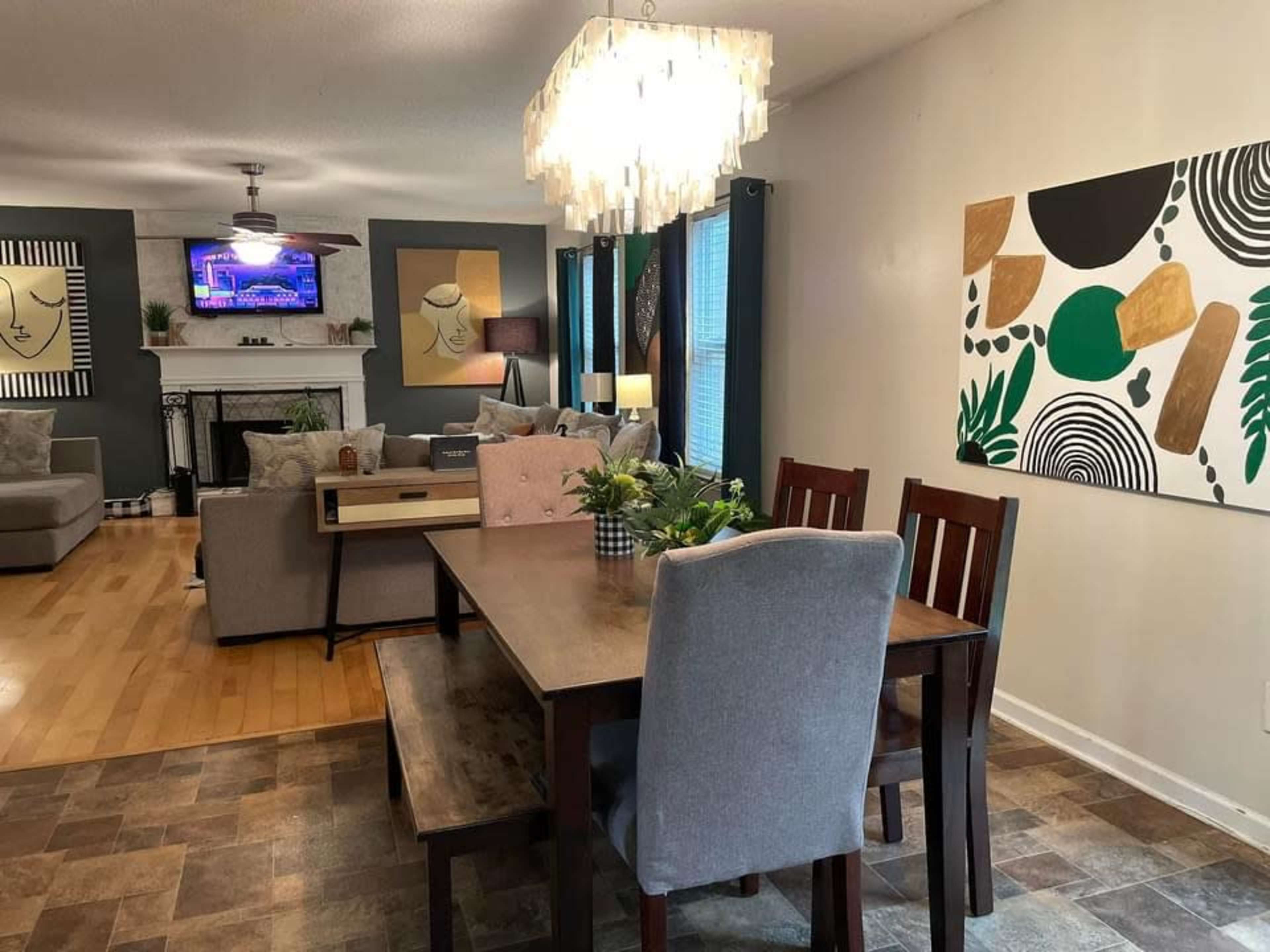 A dining area with a wooden table and chairs, a decorative chandelier, and a living room visible in the background featuring a fireplace and a TV.
