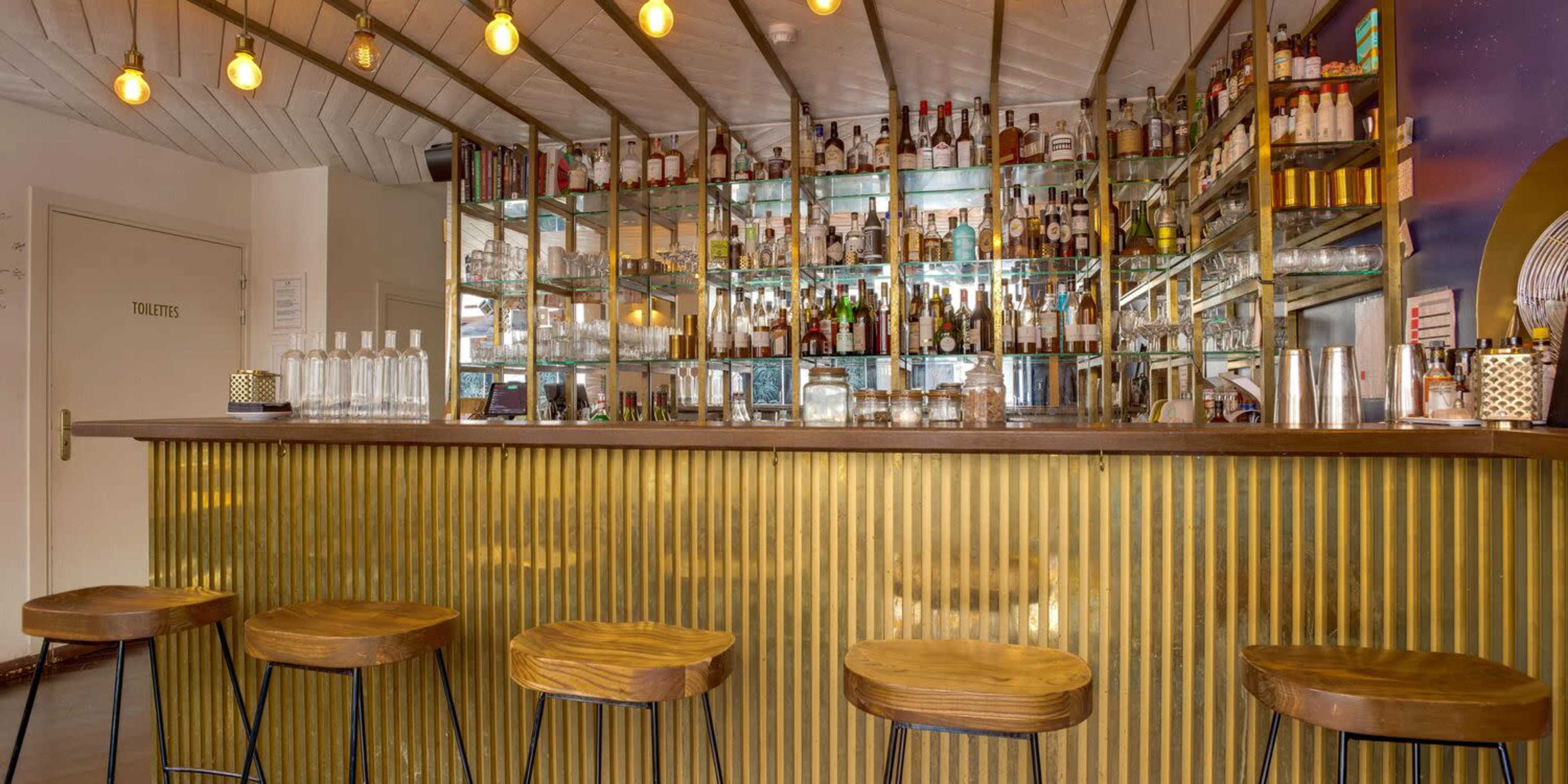 A modern bar with a brass-colored countertop, wooden stools, and a mirrored backlit shelf displaying various bottles.