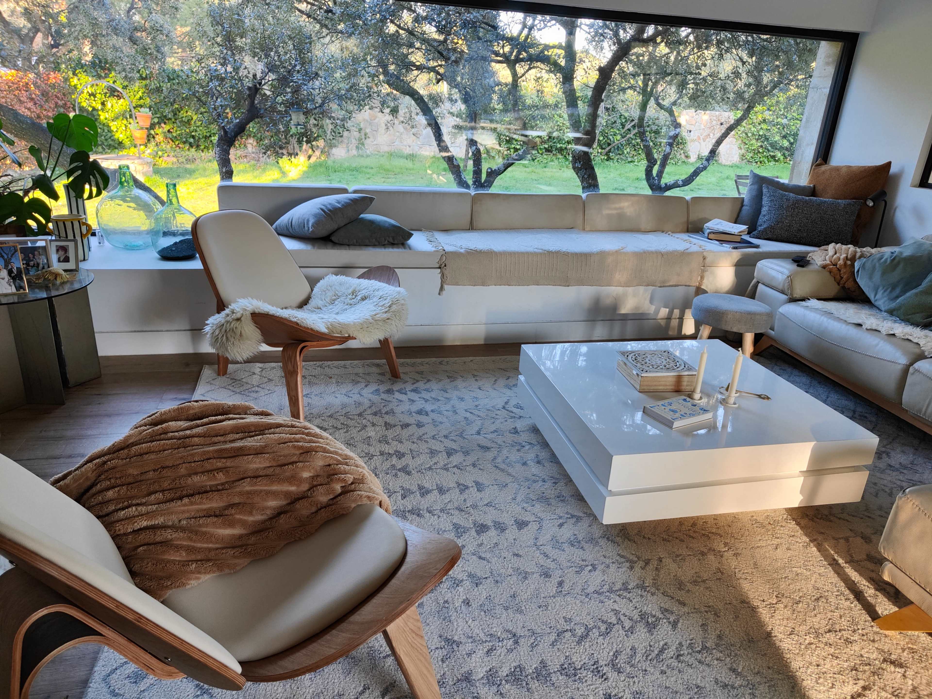 A modern living room features a light-colored sofa, a white coffee table, an armchair, and a large window overlooking a green landscape.