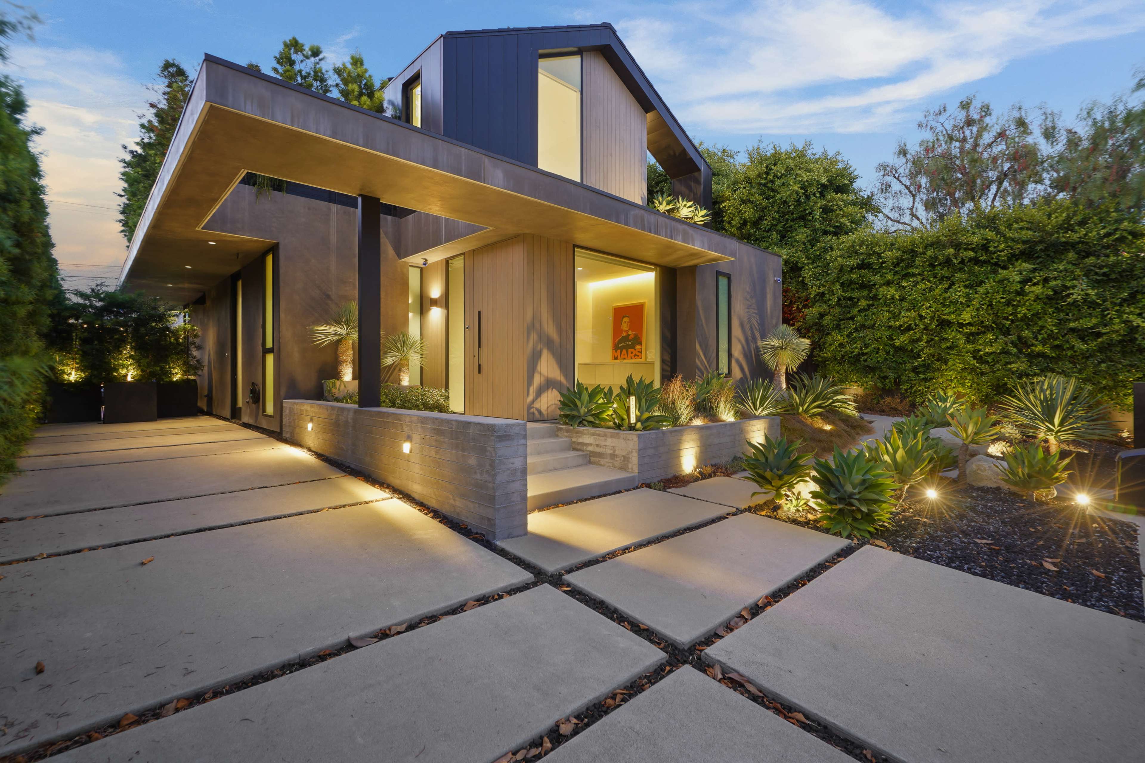 The image shows a modern house with a distinctive architectural design, featuring a landscaped path and outdoor lighting.
