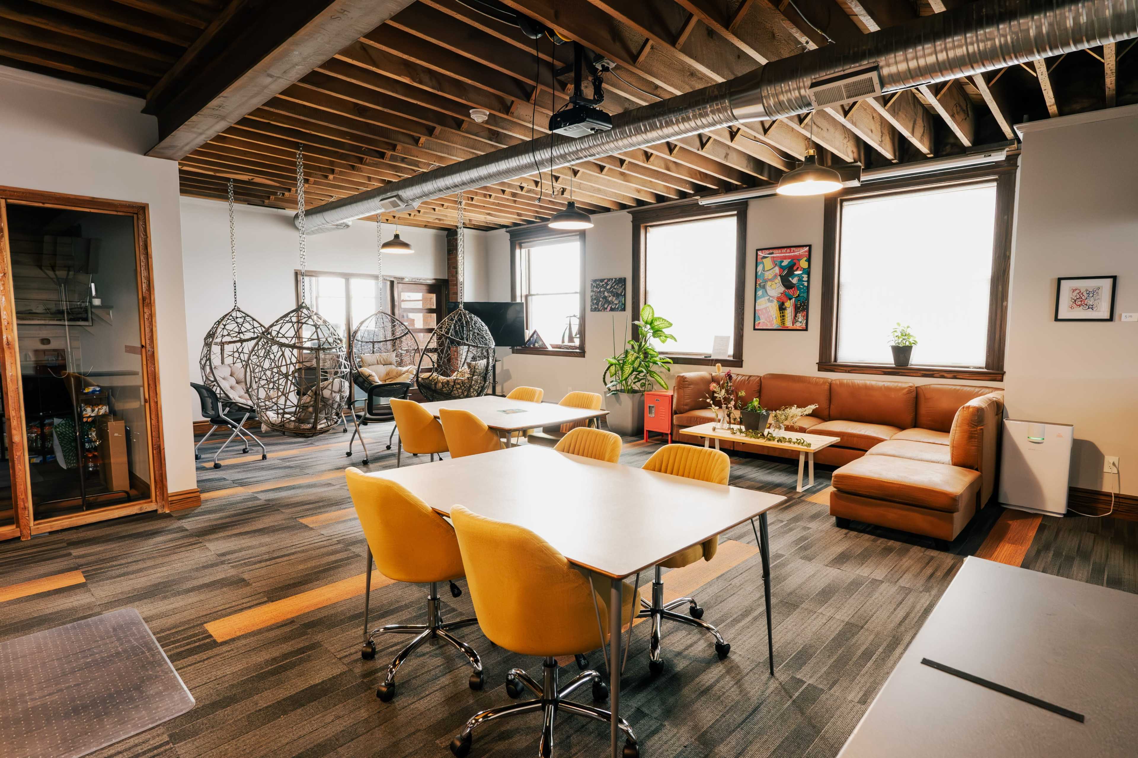 A modern office space featuring two hanging chairs, multiple work tables with yellow chairs, and a lounge area with a sectional sofa.