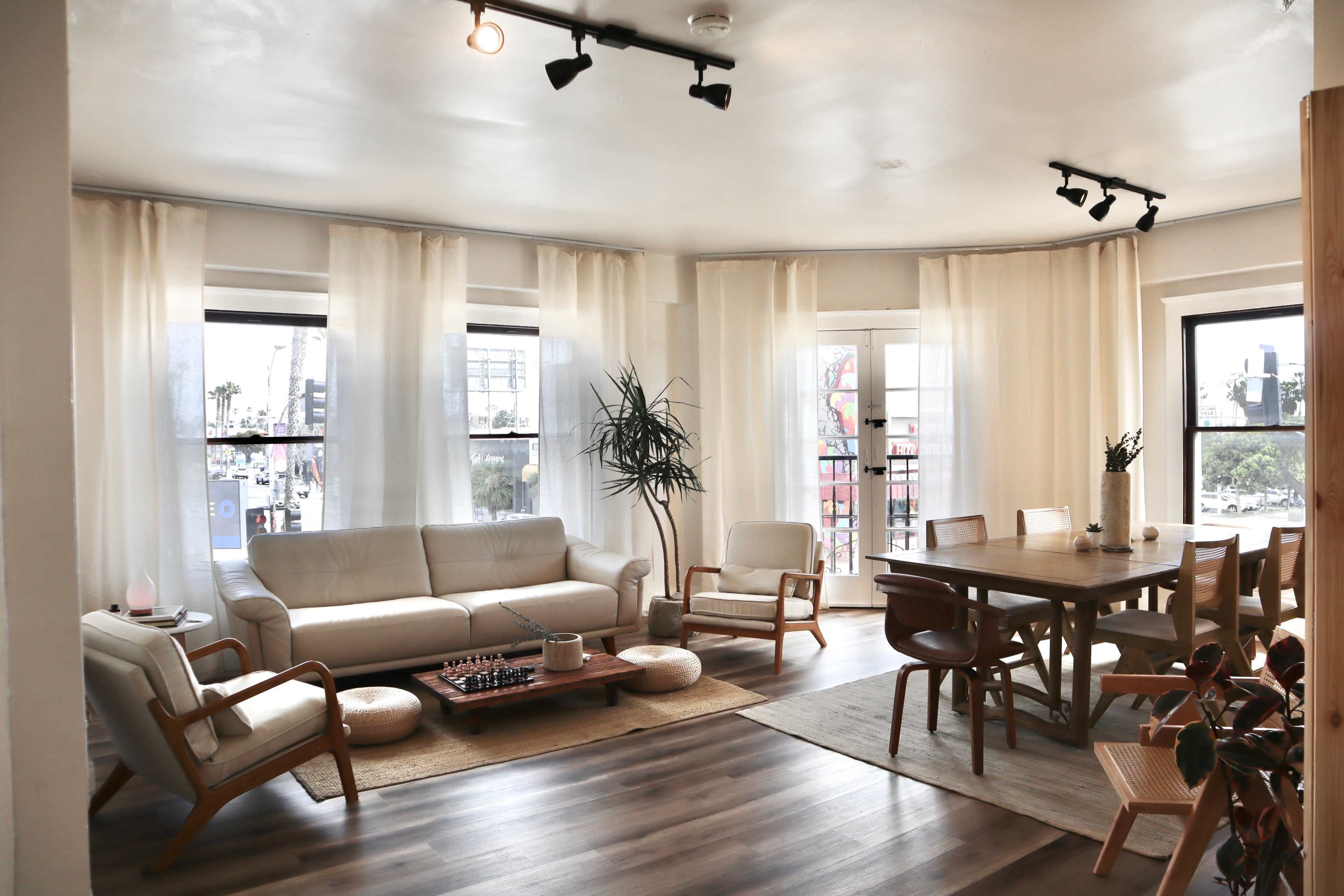 The image shows a spacious, well-lit living room featuring a white leather sofa, wooden dining table, and large windows with sheer curtains.