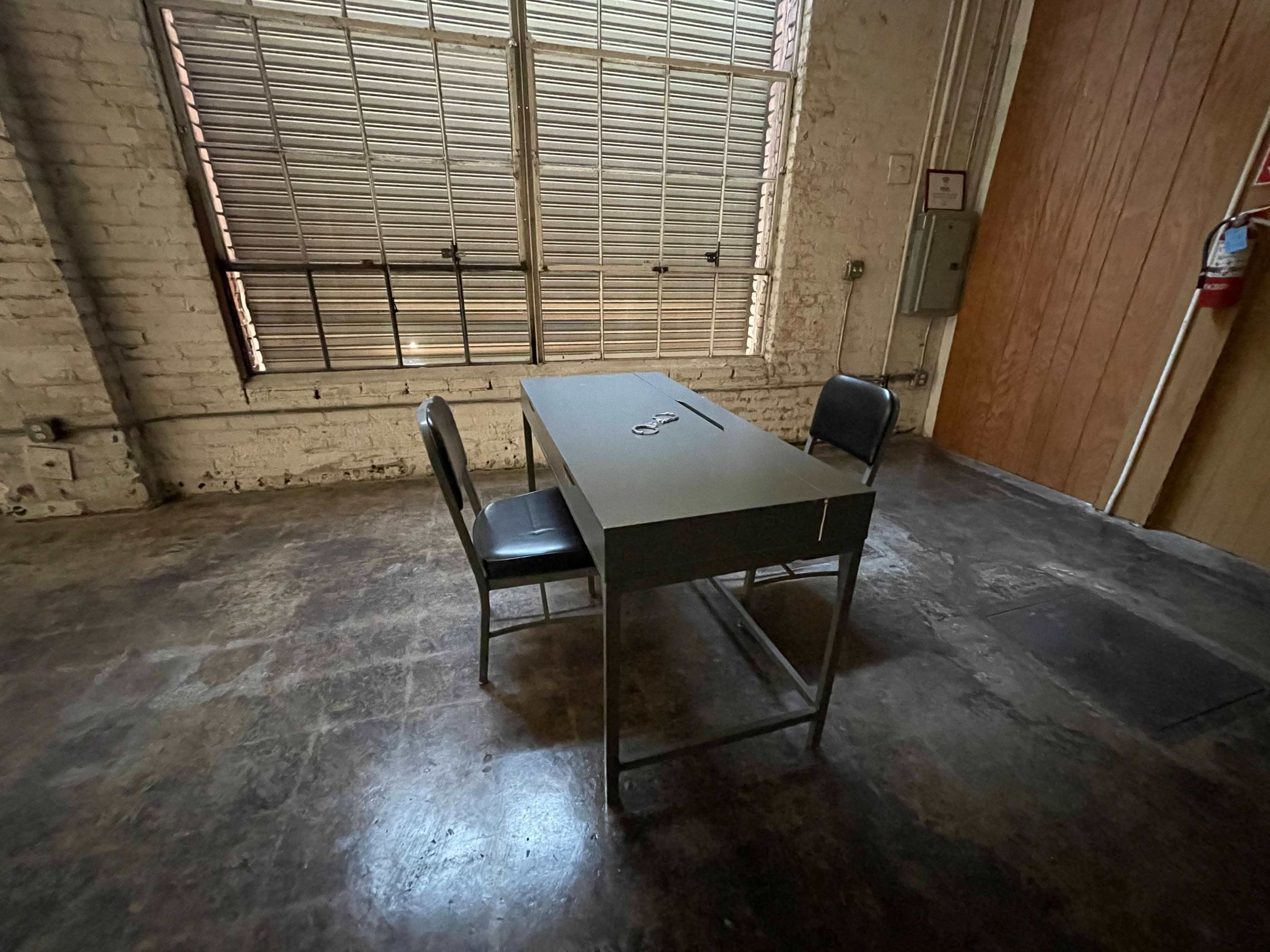 The image shows a simple table with two chairs in an empty room with exposed brick walls and a window featuring horizontal blinds.