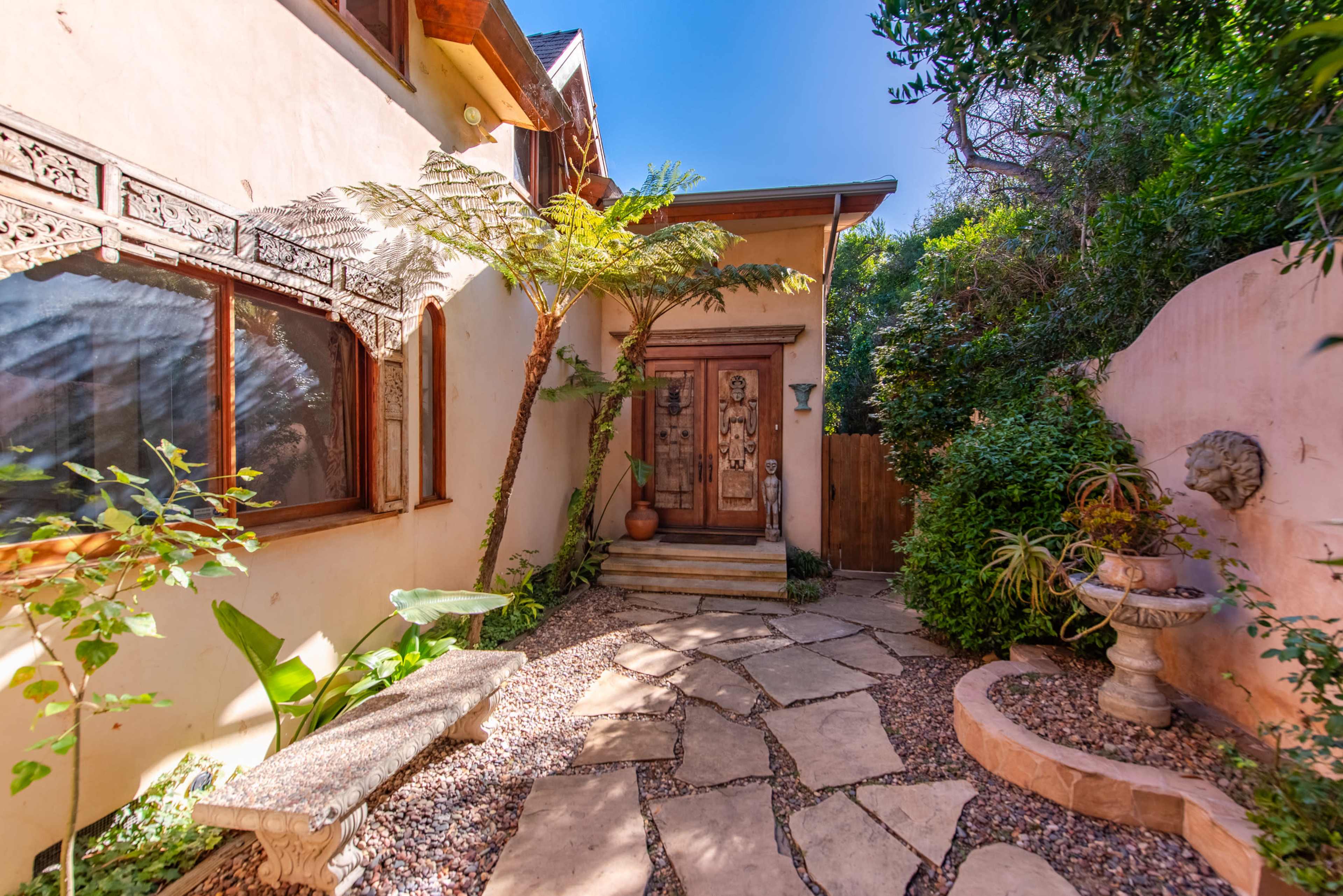 A stone pathway leading to a wooden front door, surrounded by lush greenery and a decorative stone bench in a garden setting.