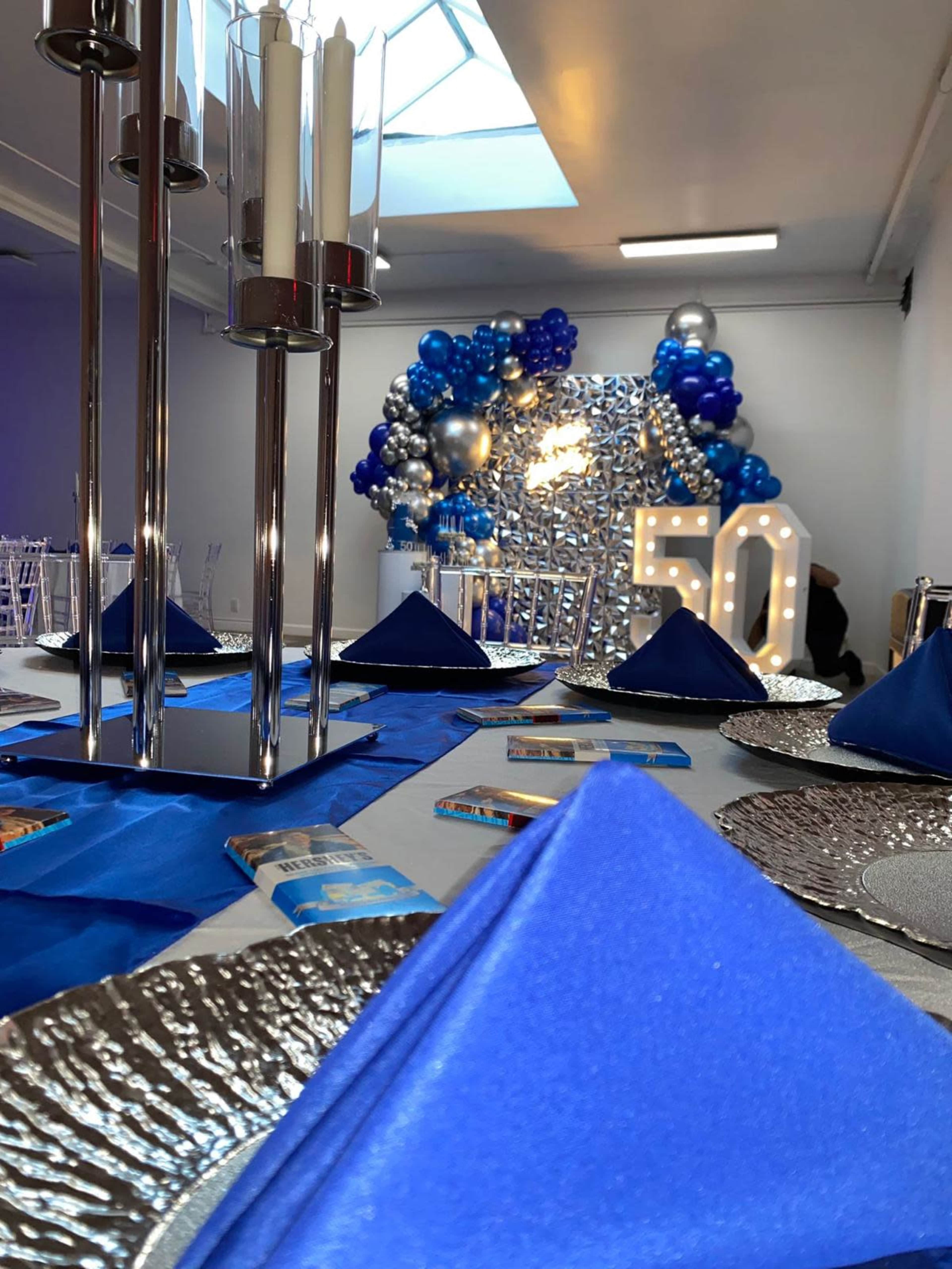 A decorated event space features a table set with blue napkins and silver chargers, while a backdrop of balloons and a large illuminated "50" is visible in the background.