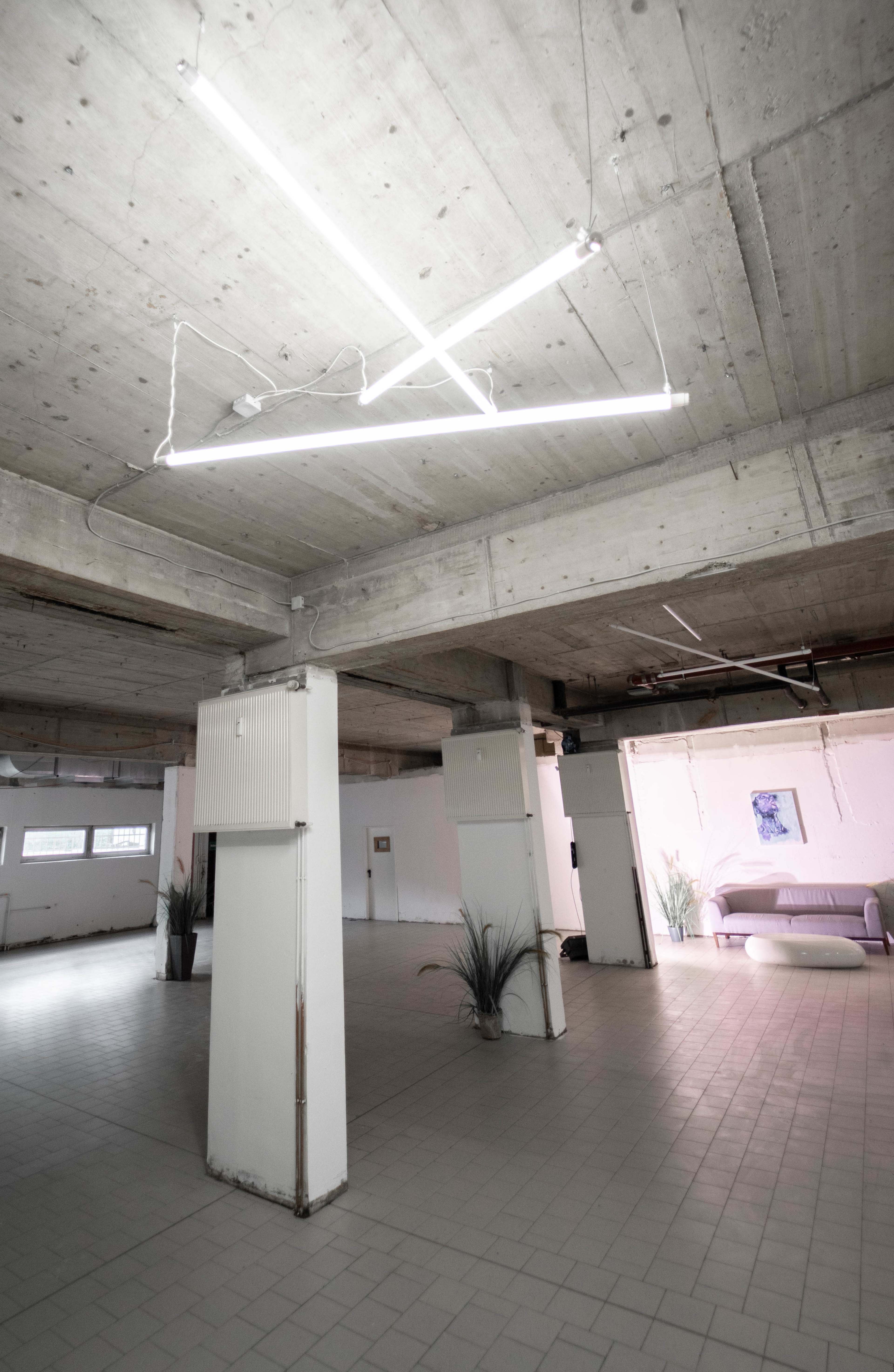 An industrial-style interior space with concrete ceilings, fluorescent lighting, and minimal furnishings, including a sofa in the background.