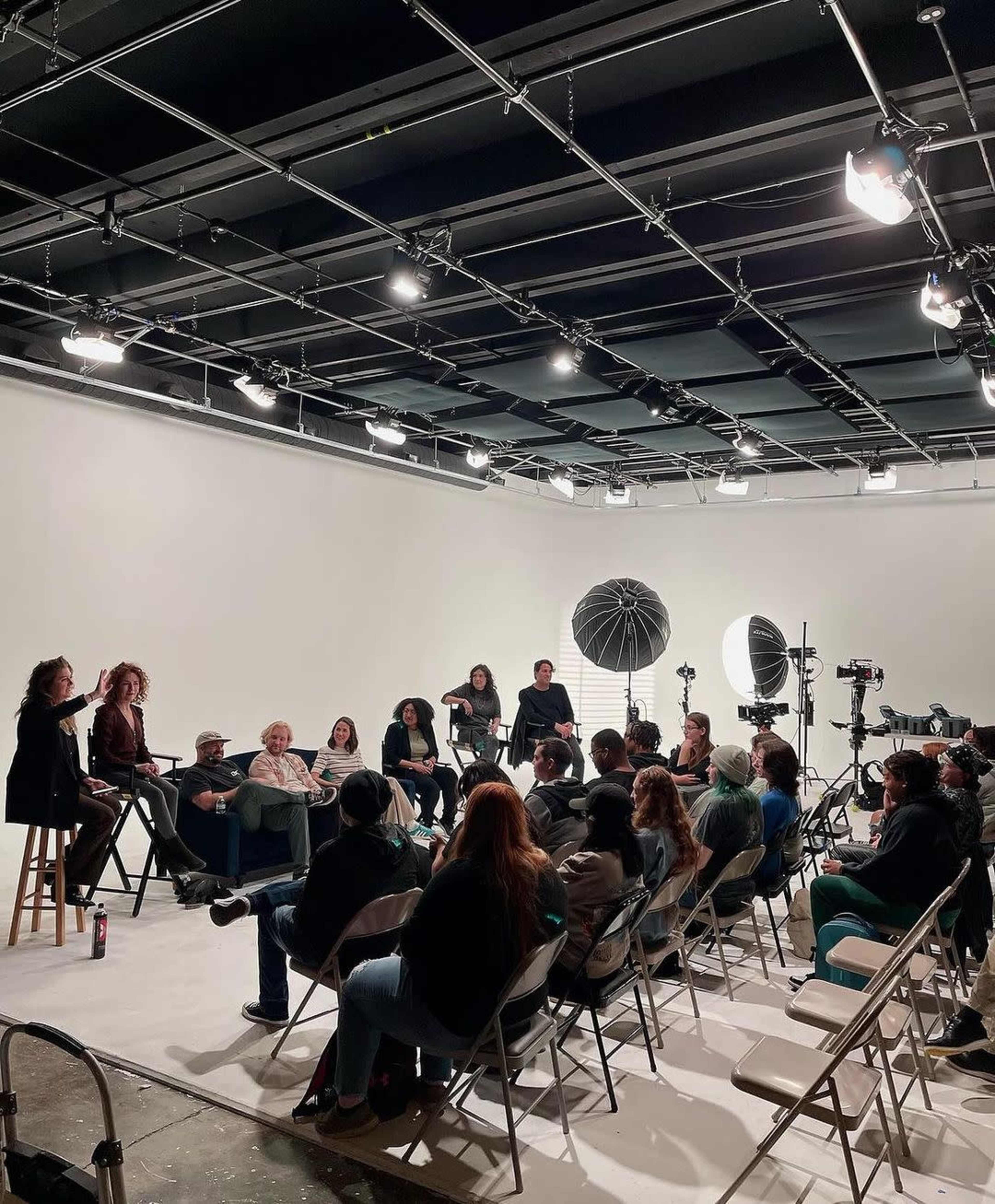 A group of people is seated in a studio, facing a panel discussion on a small stage with lighting equipment and cameras set up around them.