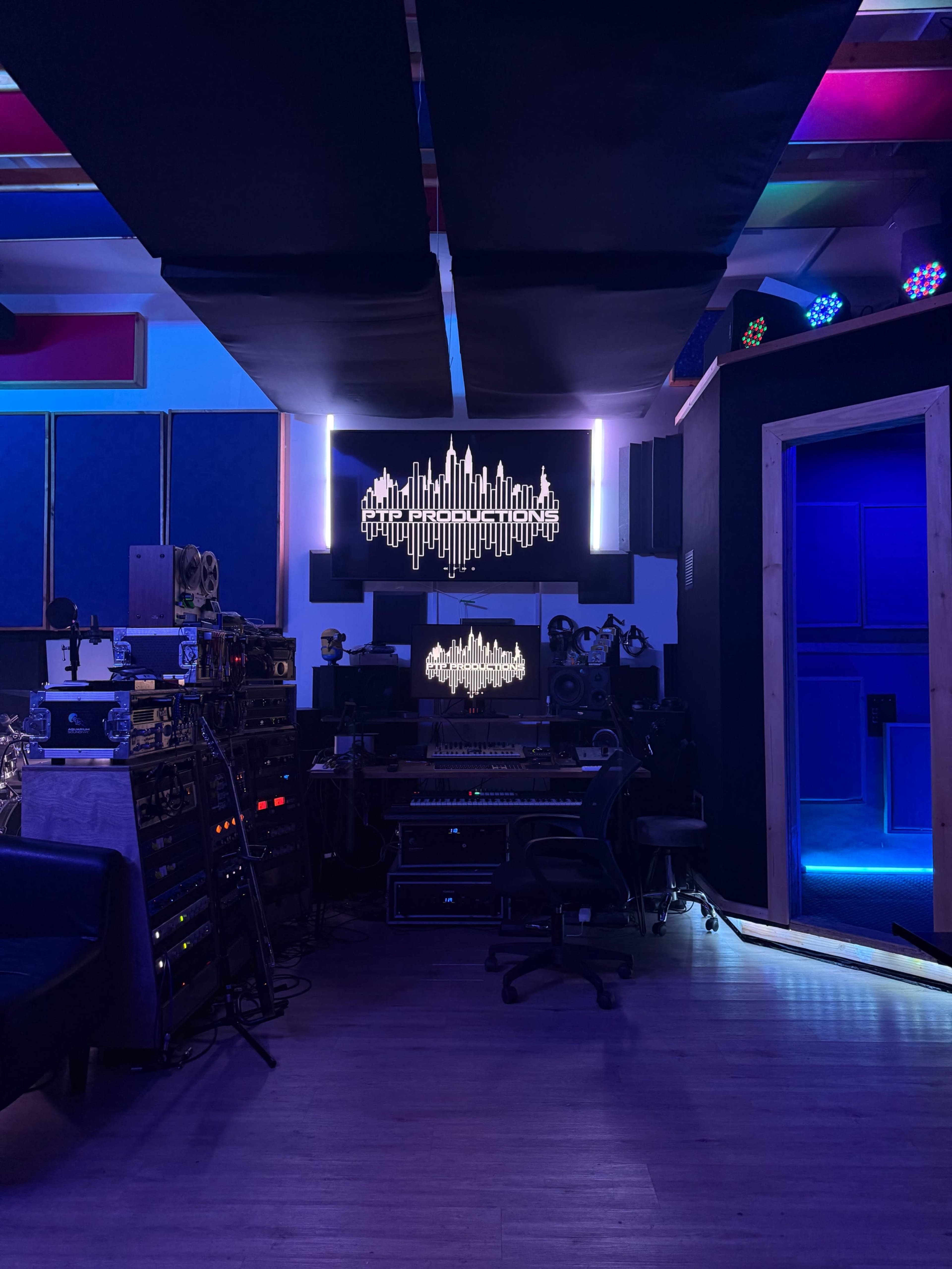 The image shows a music studio with various audio equipment, a computer setup, and soundproofing panels on the walls.