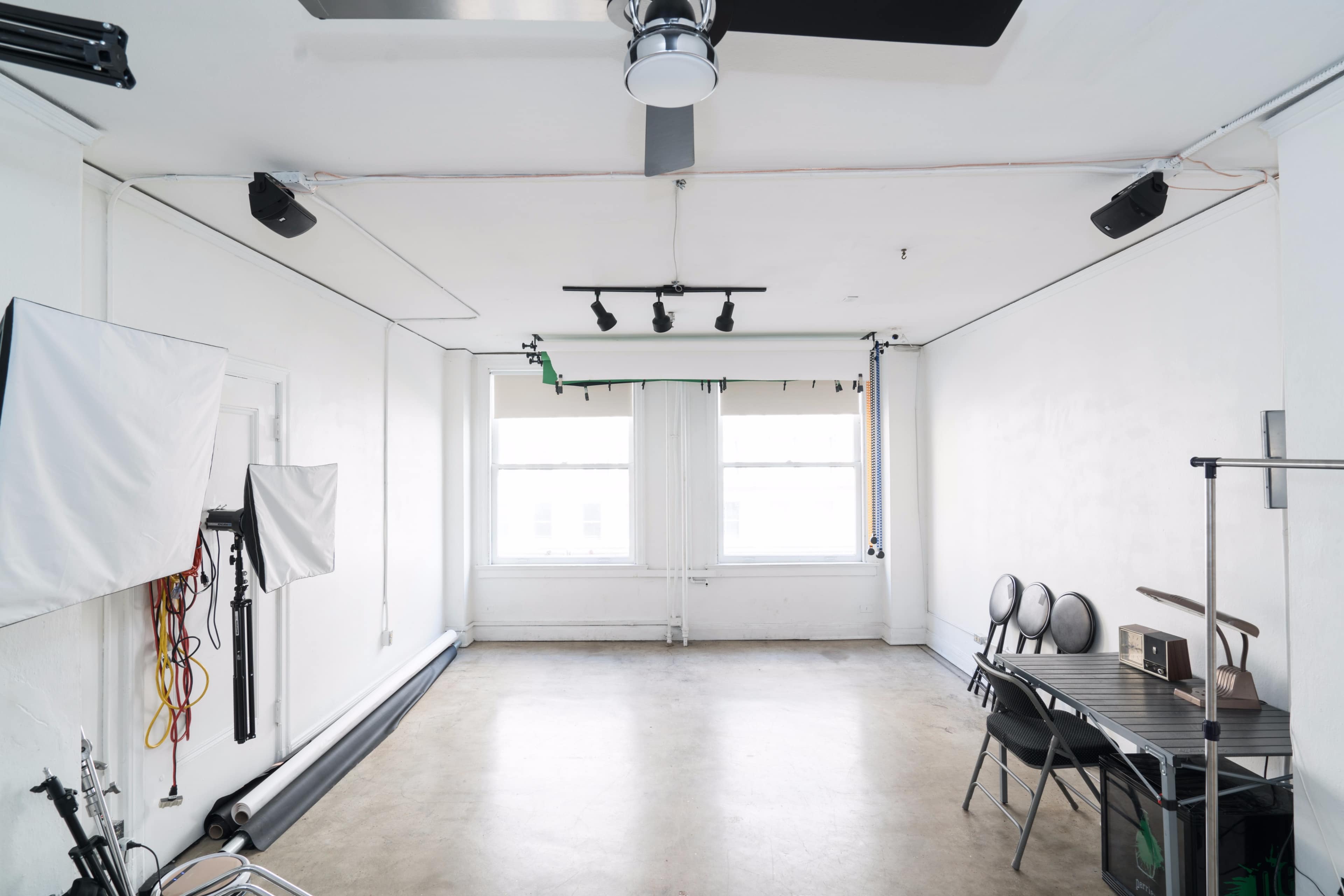 A spacious, empty studio with concrete flooring, white walls, two large windows, and a setup for photography that includes light stands and a folding table with chairs.