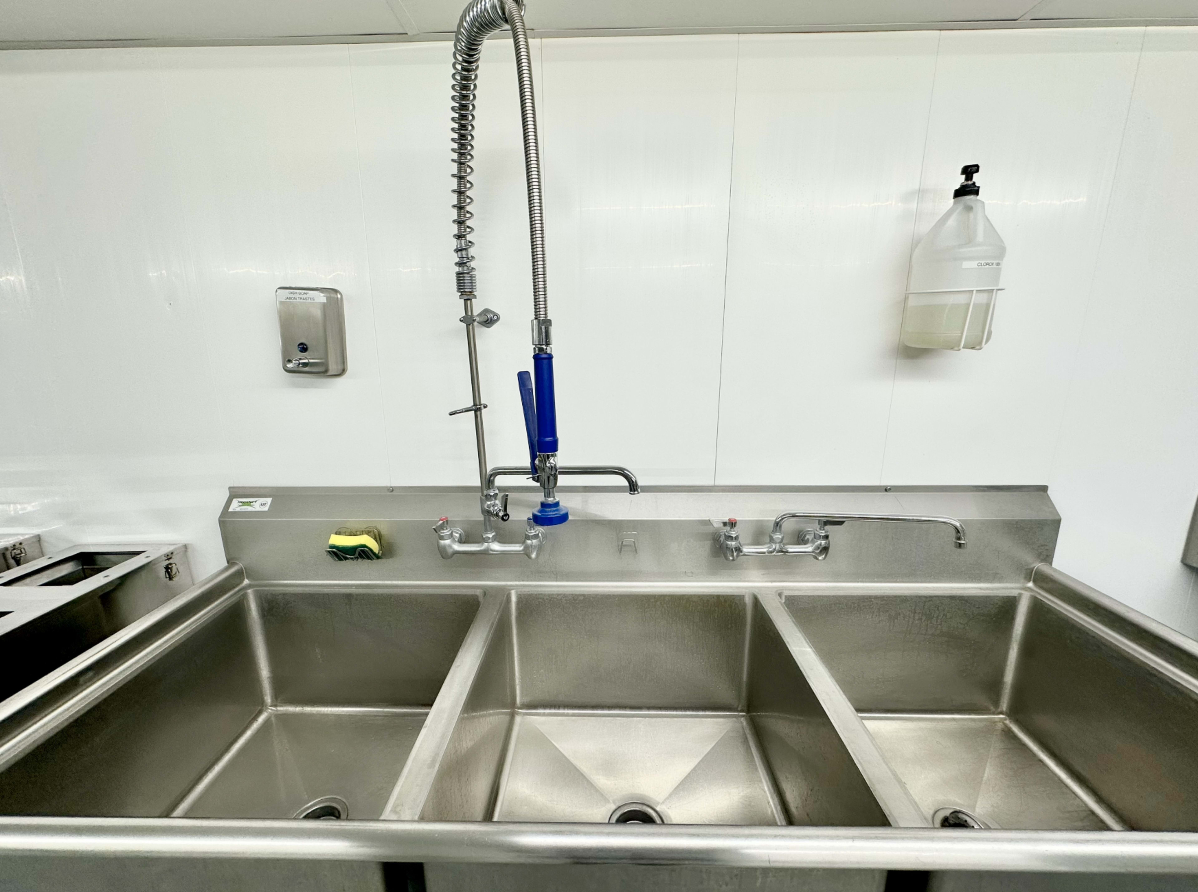 The image shows a stainless steel sink with three compartments, a faucet with a sprayer attachment, and a soap dispenser mounted on the wall.