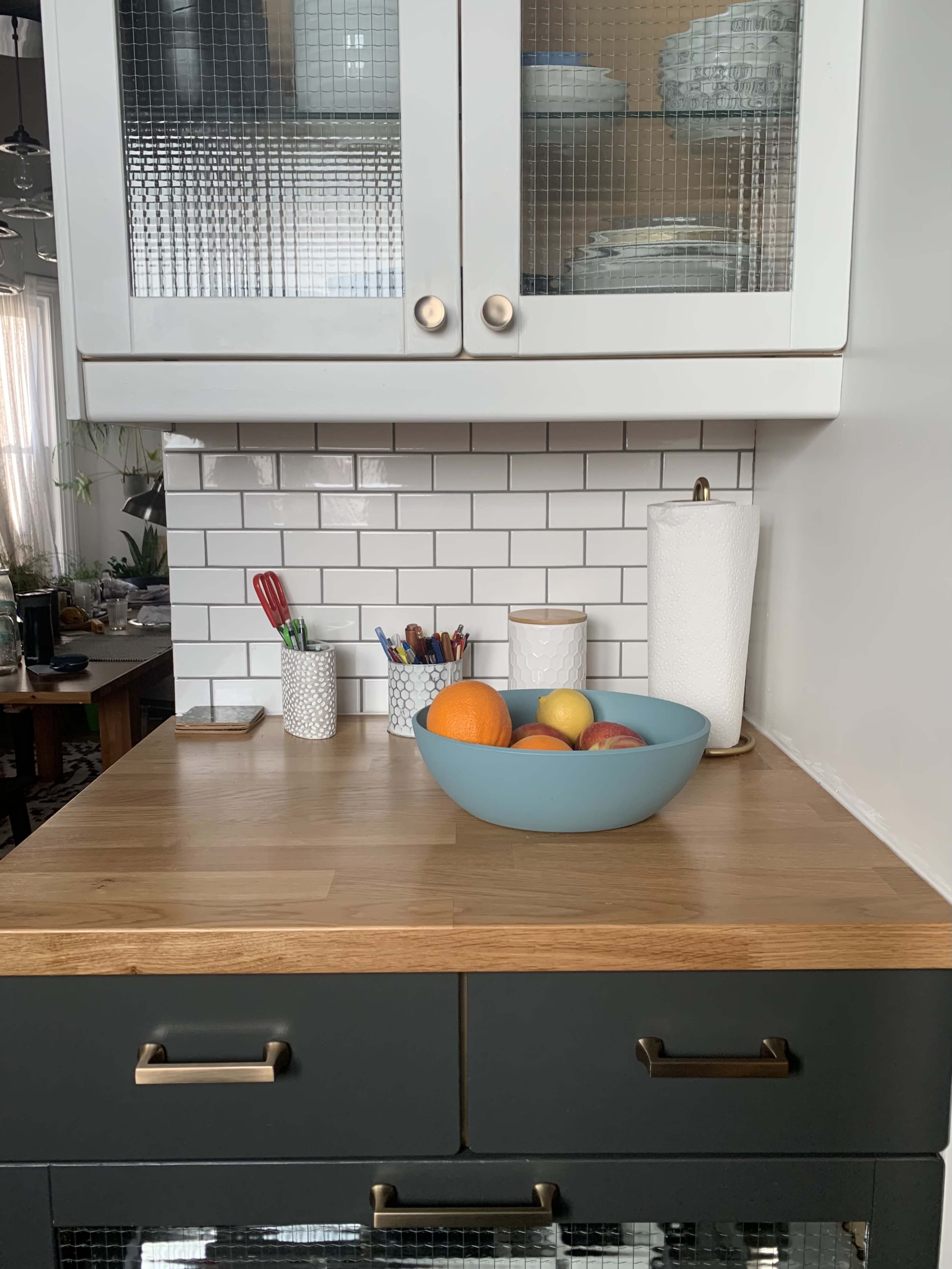 A countertop displays a blue bowl of assorted fruits, surrounded by utensils and containers on a kitchen surface.