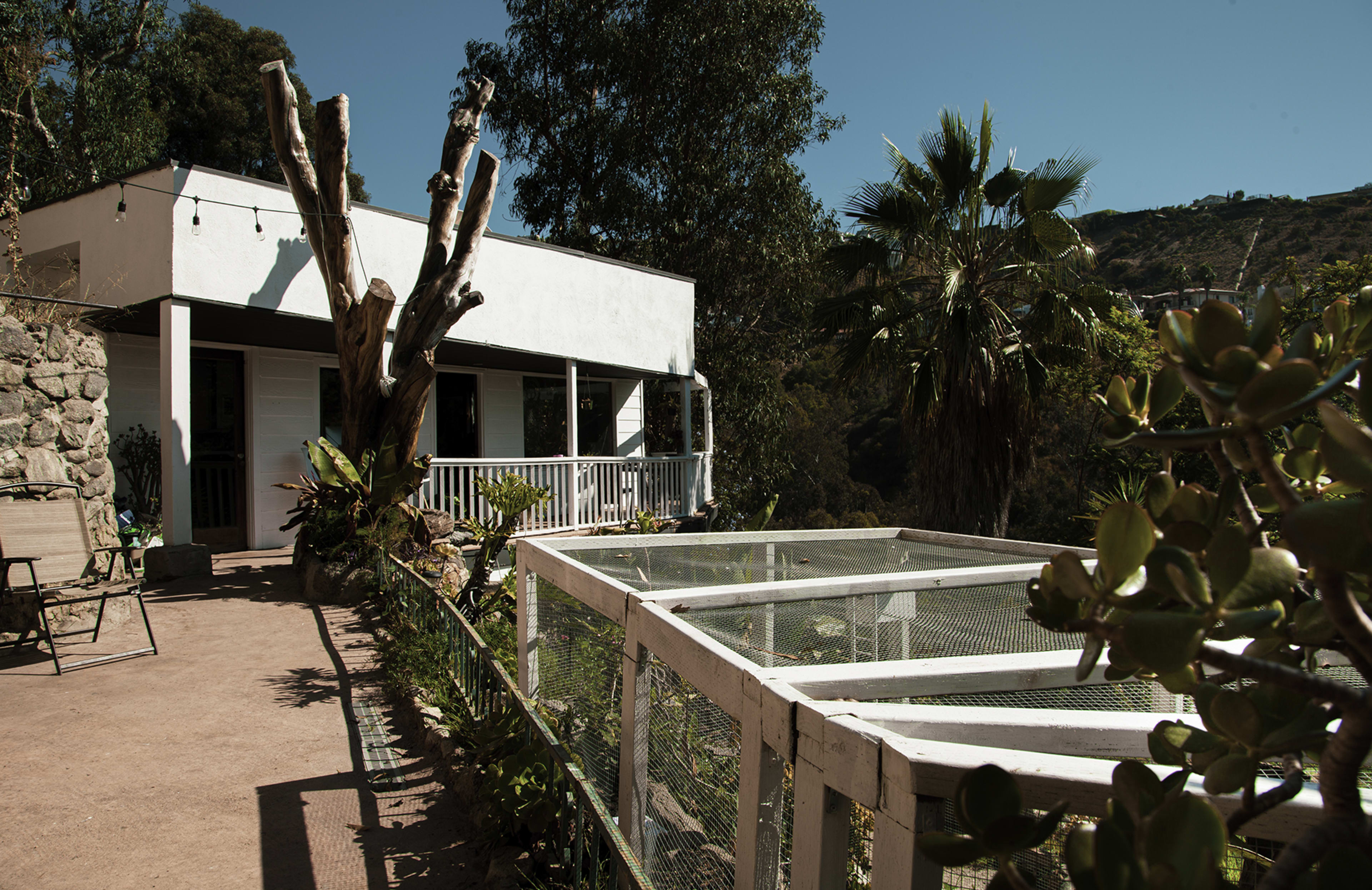A white house with a porch is surrounded by trees and plants, with a fenced garden area in the foreground.