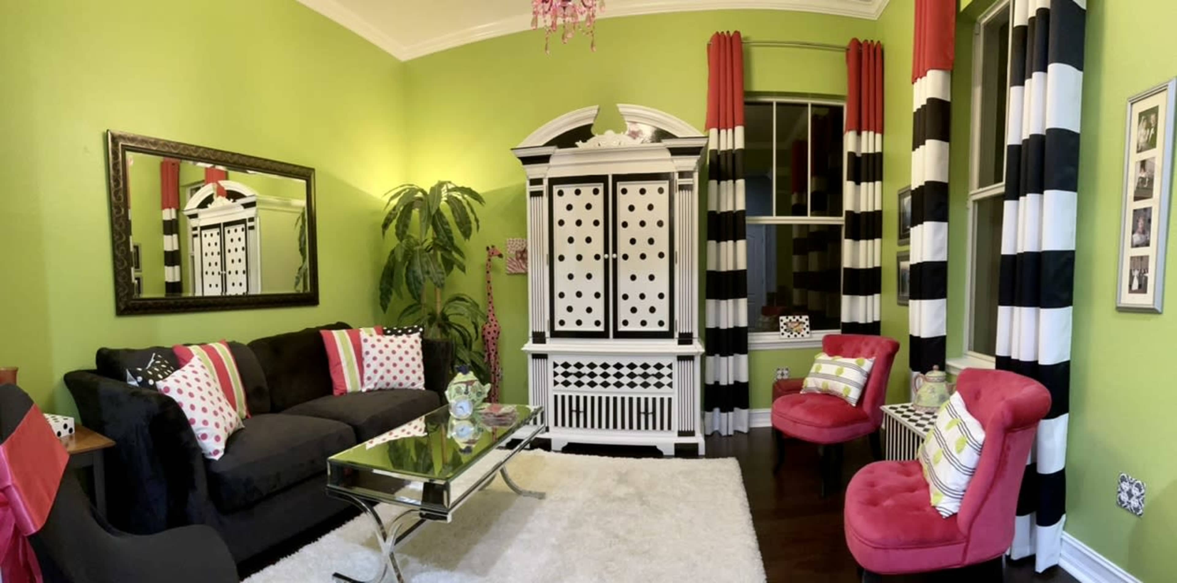 The room features a green wall, a black sofa with decorative pillows, a silver coffee table, striped black and white curtains, and a furniture piece with polka dots.