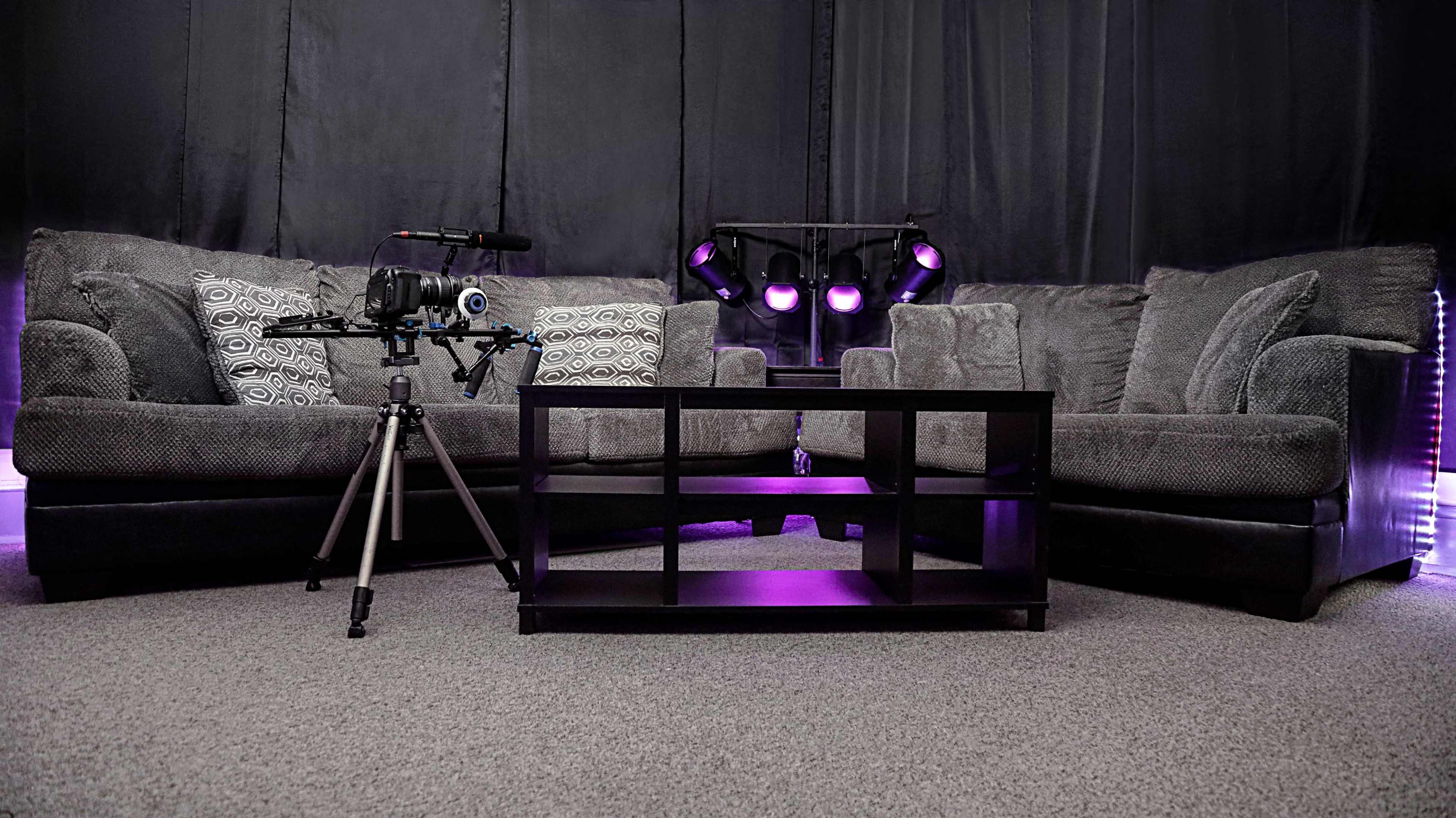 A cozy living room setup with a gray couch, a black coffee table, and a video camera mounted on a tripod in front of a backdrop with purple lighting.