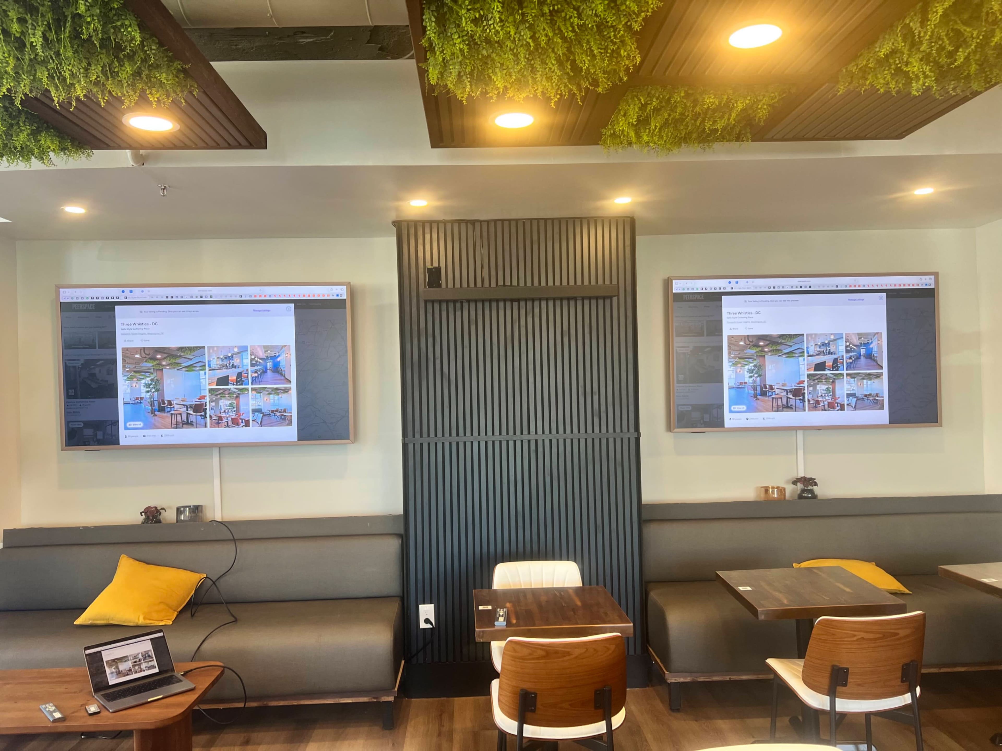 The image shows a modern café interior with two large screens displaying images, along with comfortable seating and wooden tables.