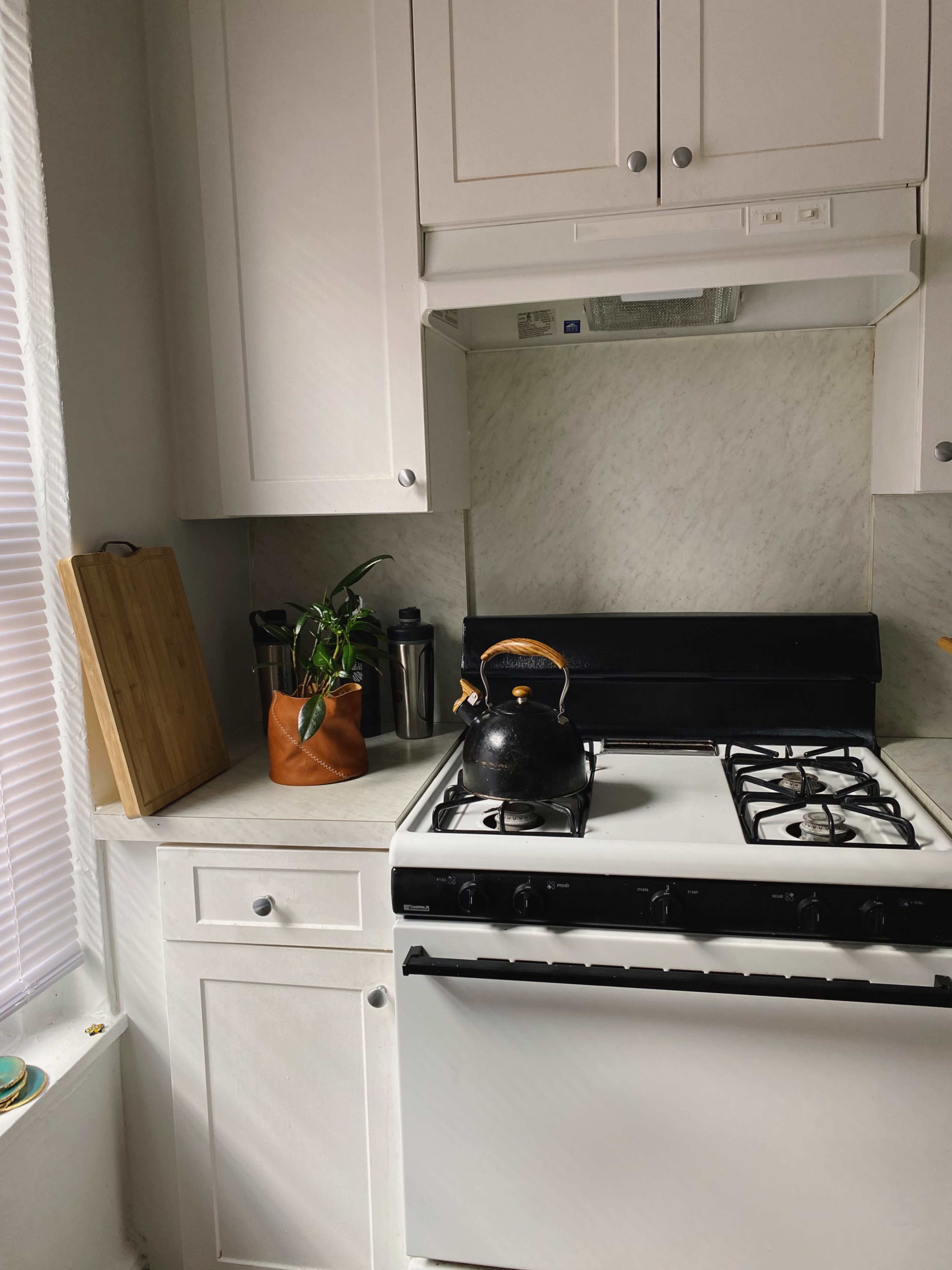 A black kettle sits on a gas stove in a modern kitchen with white cabinetry and a marble backsplash.