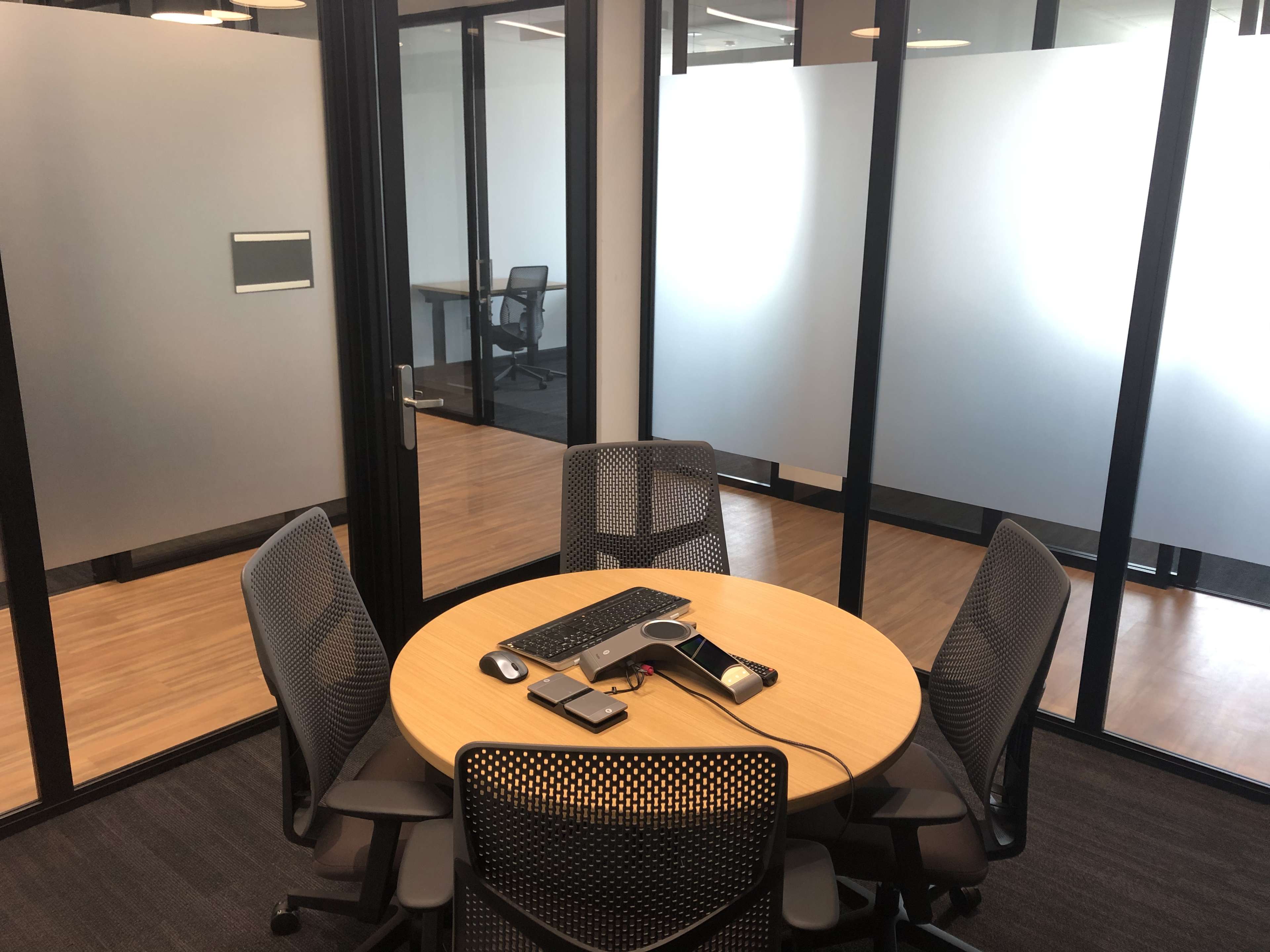 A round table with a computer and phone sits in a glass-walled meeting room featuring four mesh chairs and wooden flooring.