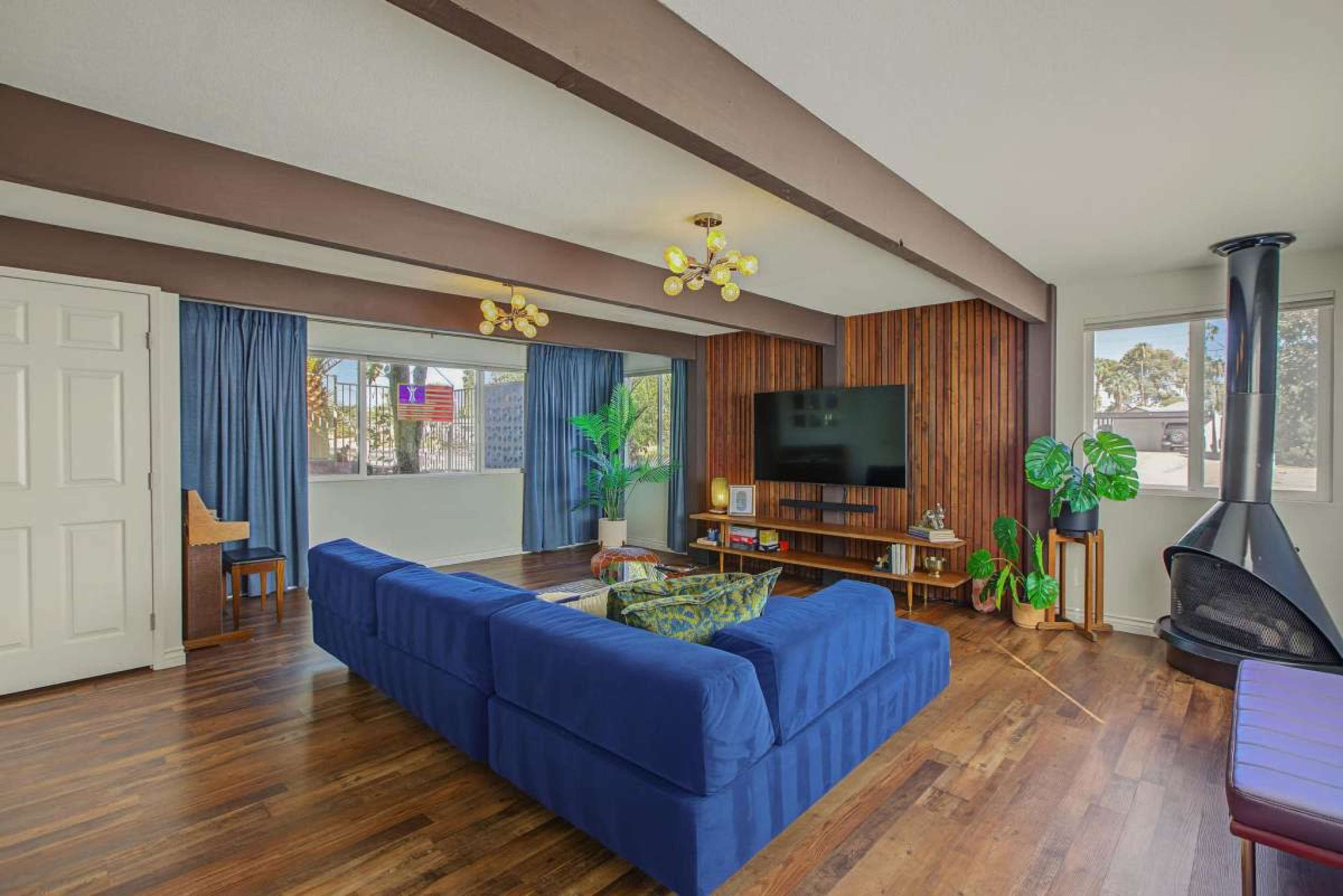 The image shows a living room with a blue sectional sofa, a wall-mounted television, and large windows covered with blue curtains.